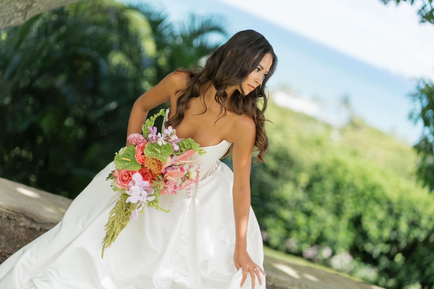 Bride in strapless white gown holding colorful bouquet of tropical flowers, posing outdoors with ocean view.