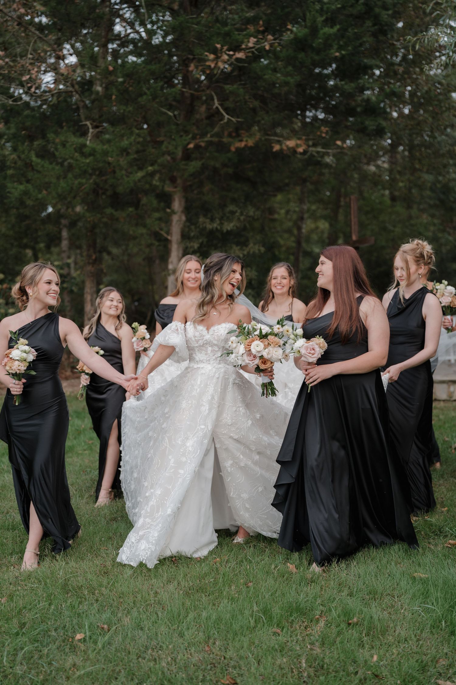 A wedding party poses playfully in a line on grass wearing black dresses with the bride in white.
