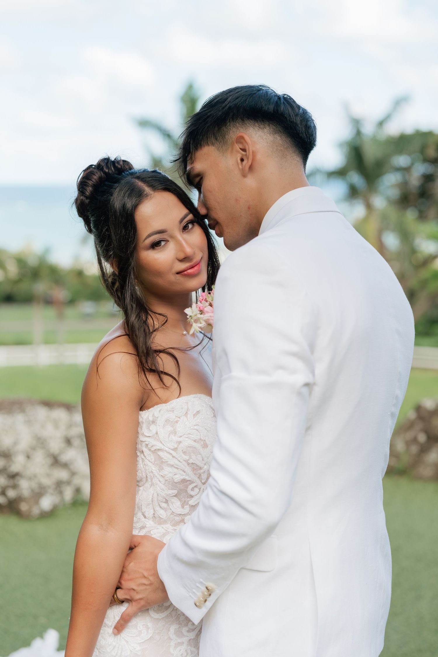 A couple in wedding attire embraces outdoors, with the groom in a white suit and bride in a lace gown, tropical scenery behind them.