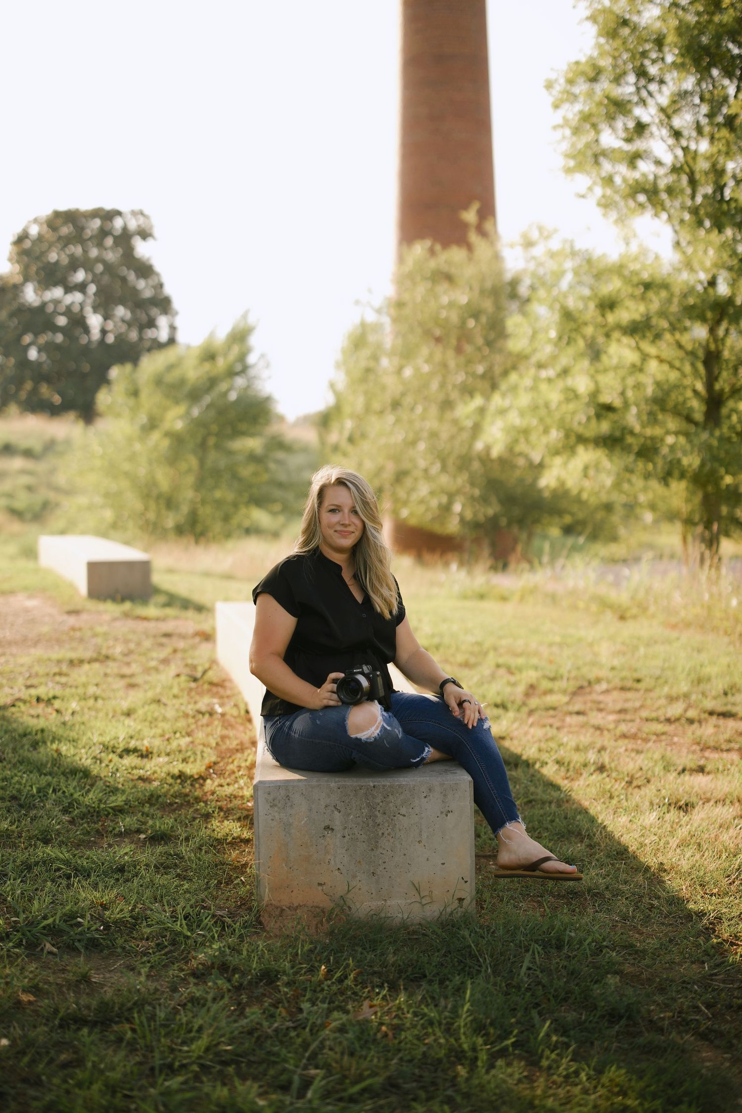 A photographer sits casually on a concrete block with a tall brick chimney in the background.