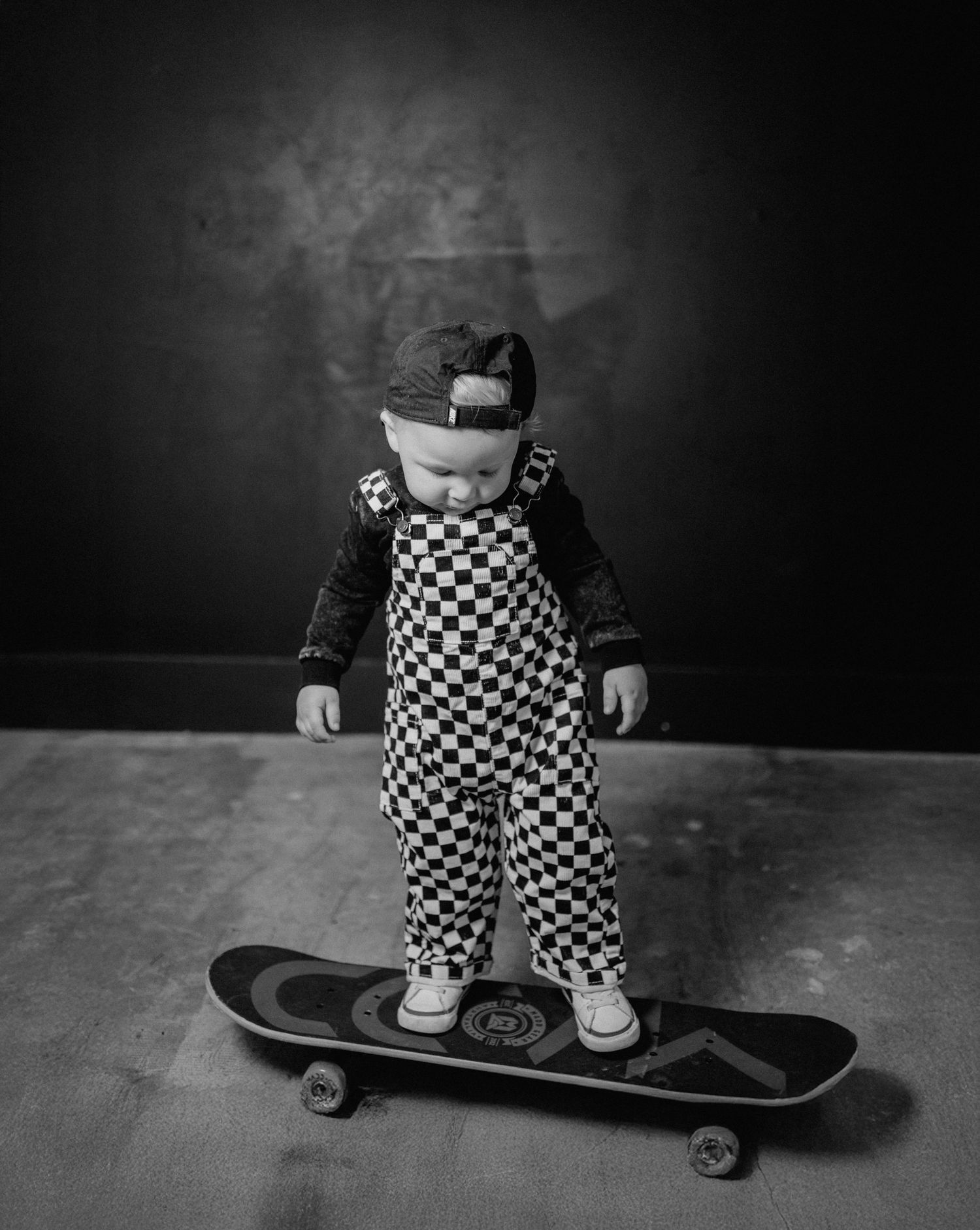 Black and white photo of a cute toddler in checkered overalls and cap standing on a skateboard.