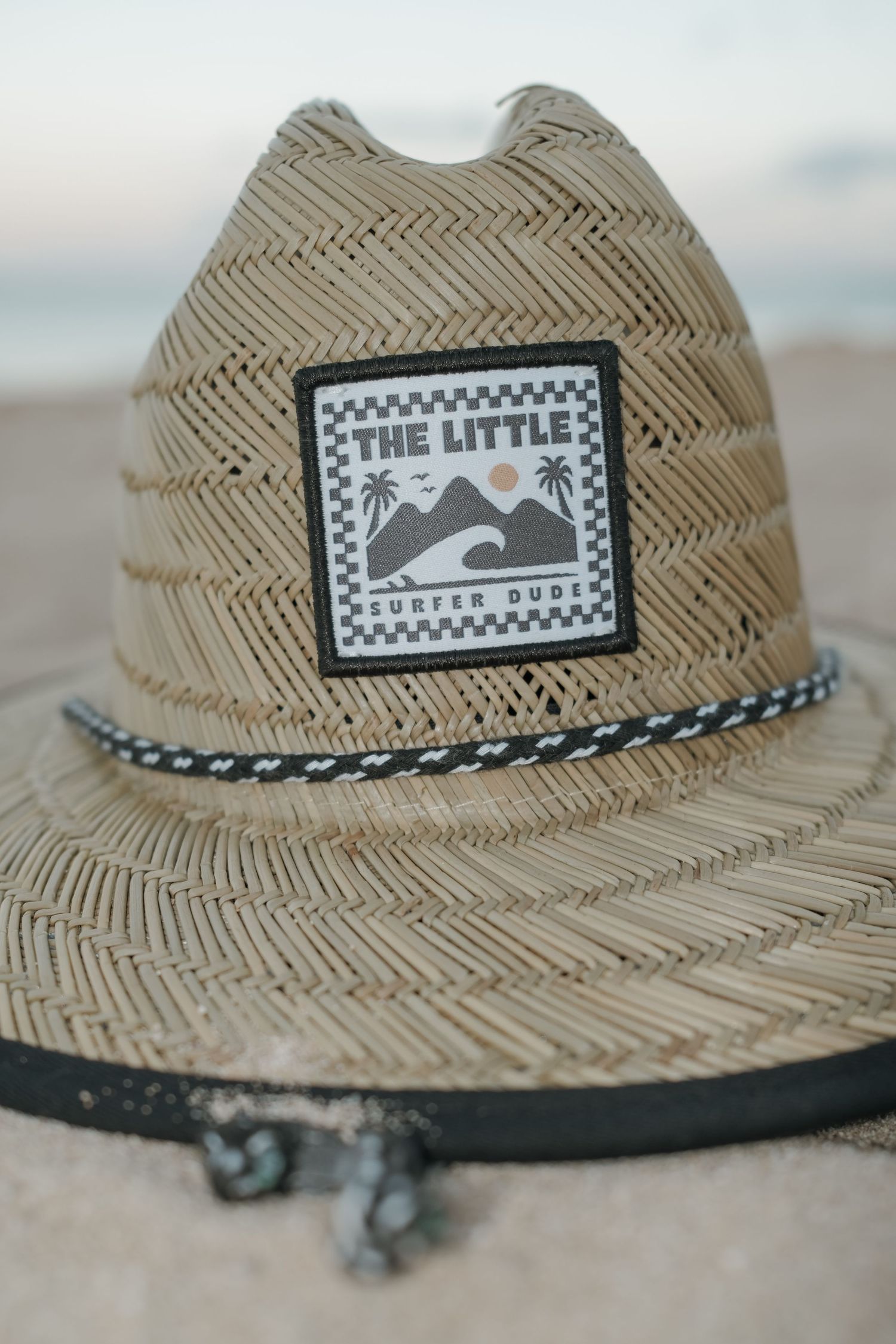 Straw lifeguard hat with The Little Surfer Dude patch on a beach.