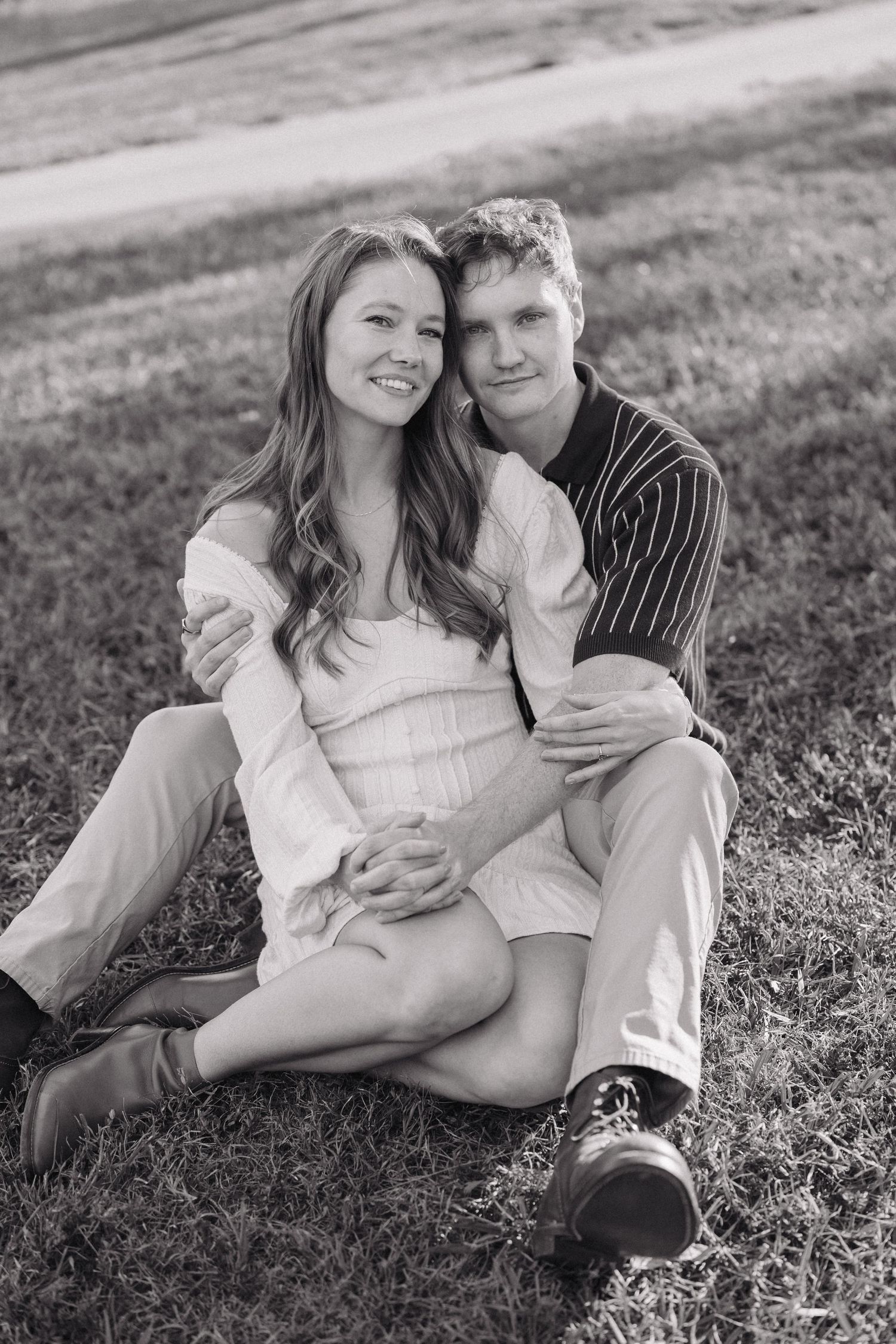 A romantic black and white portrait of a couple sitting together on grass, sharing an intimate moment outdoors.