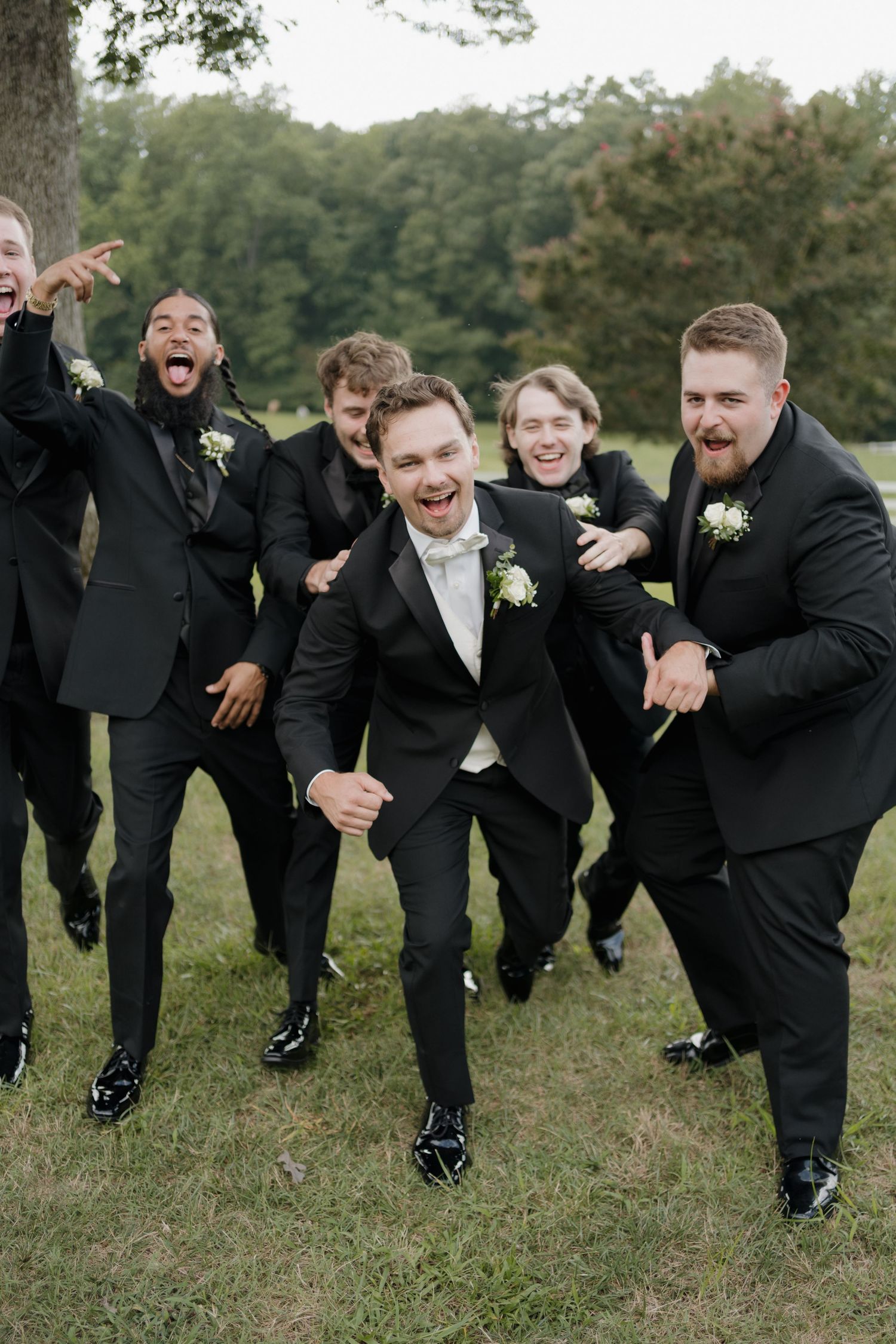 Groomsmen in black suits and boutonnieres share a joyful candid moment while laughing and jumping outdoors.