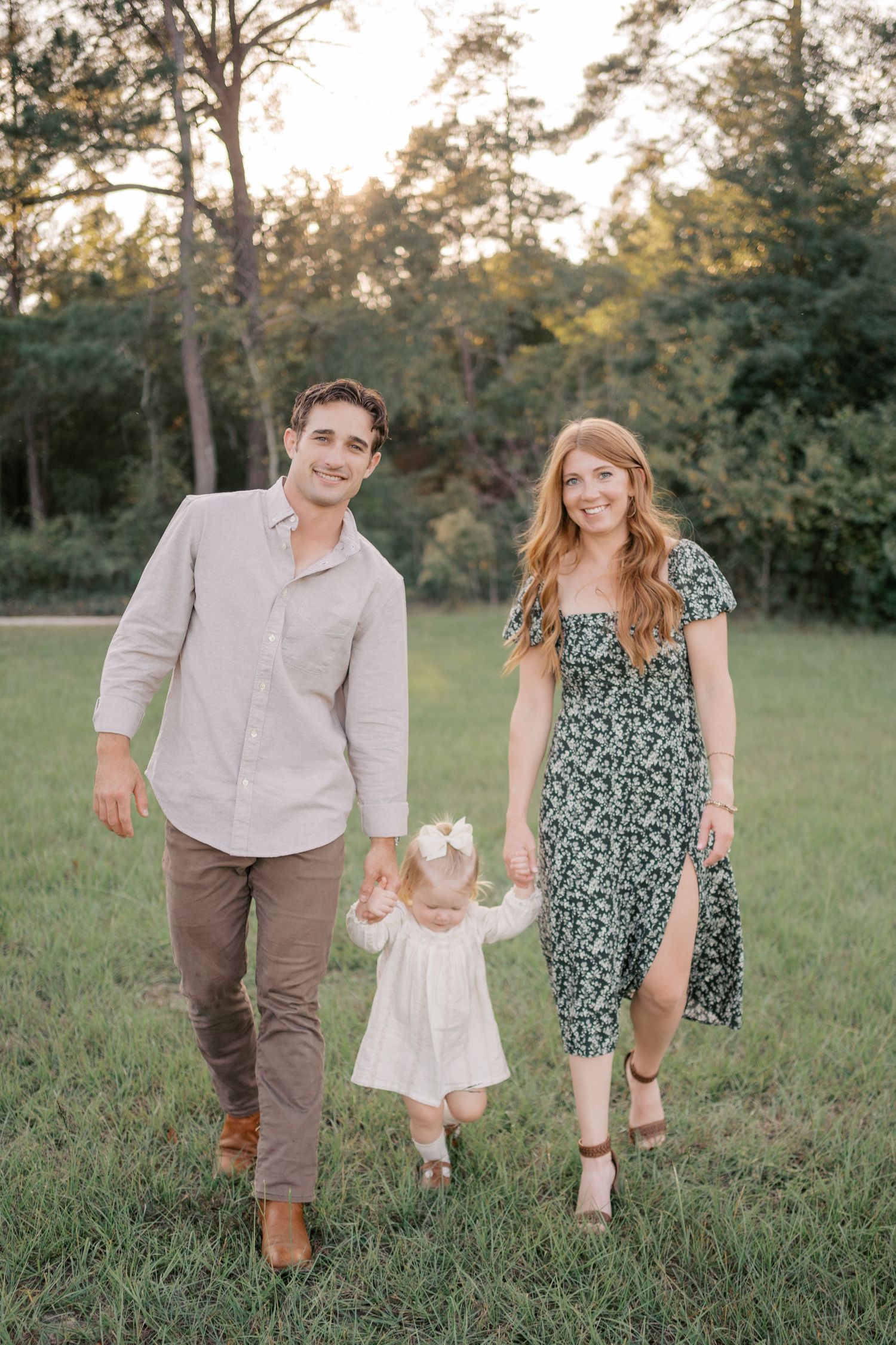 A family holding hands while walking together in a grassy field at sunset with trees in the background.