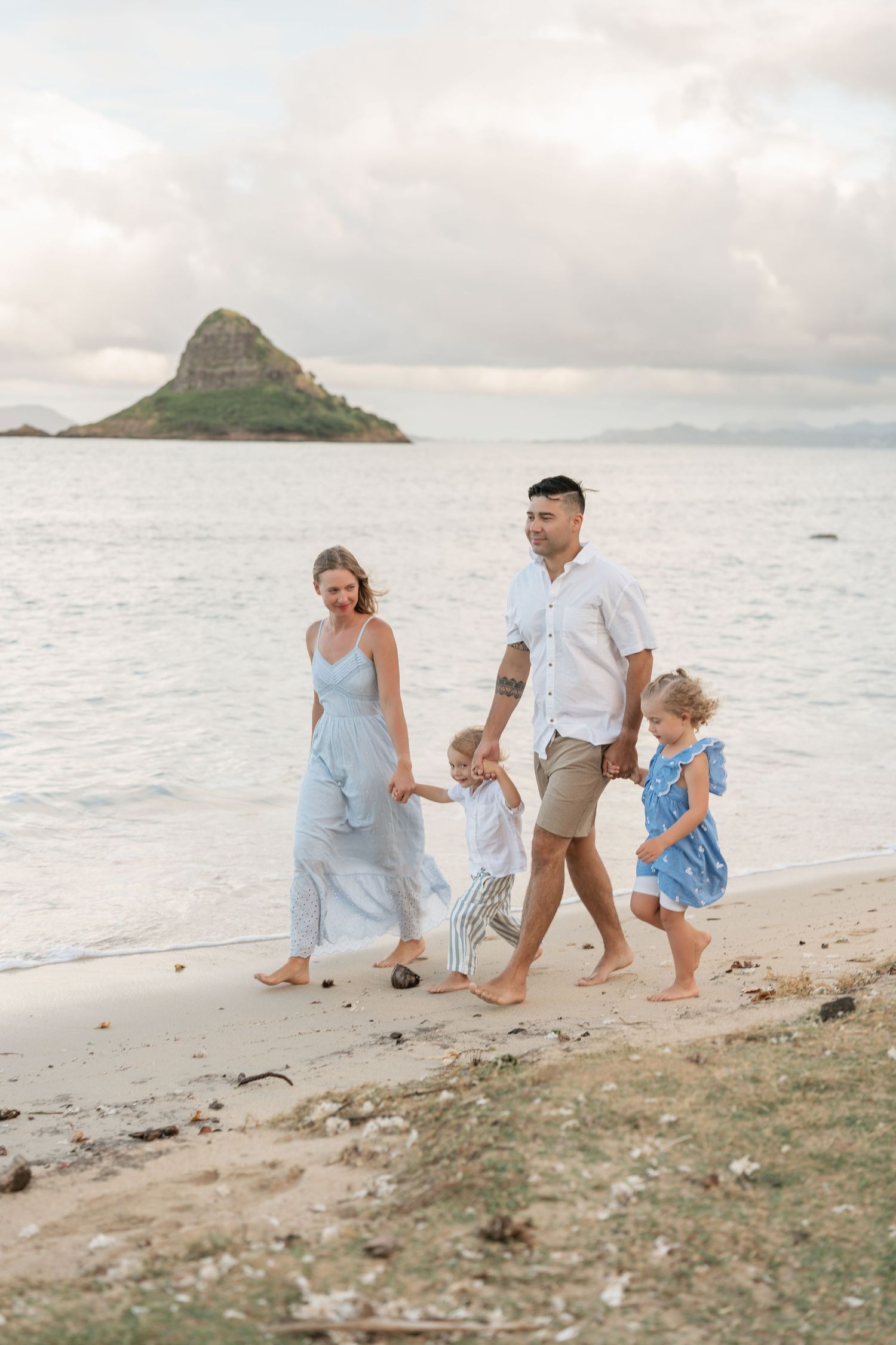 Family of four walking hand-in-hand on a Hawaiian beach with Chinaman's Hat island in the background.