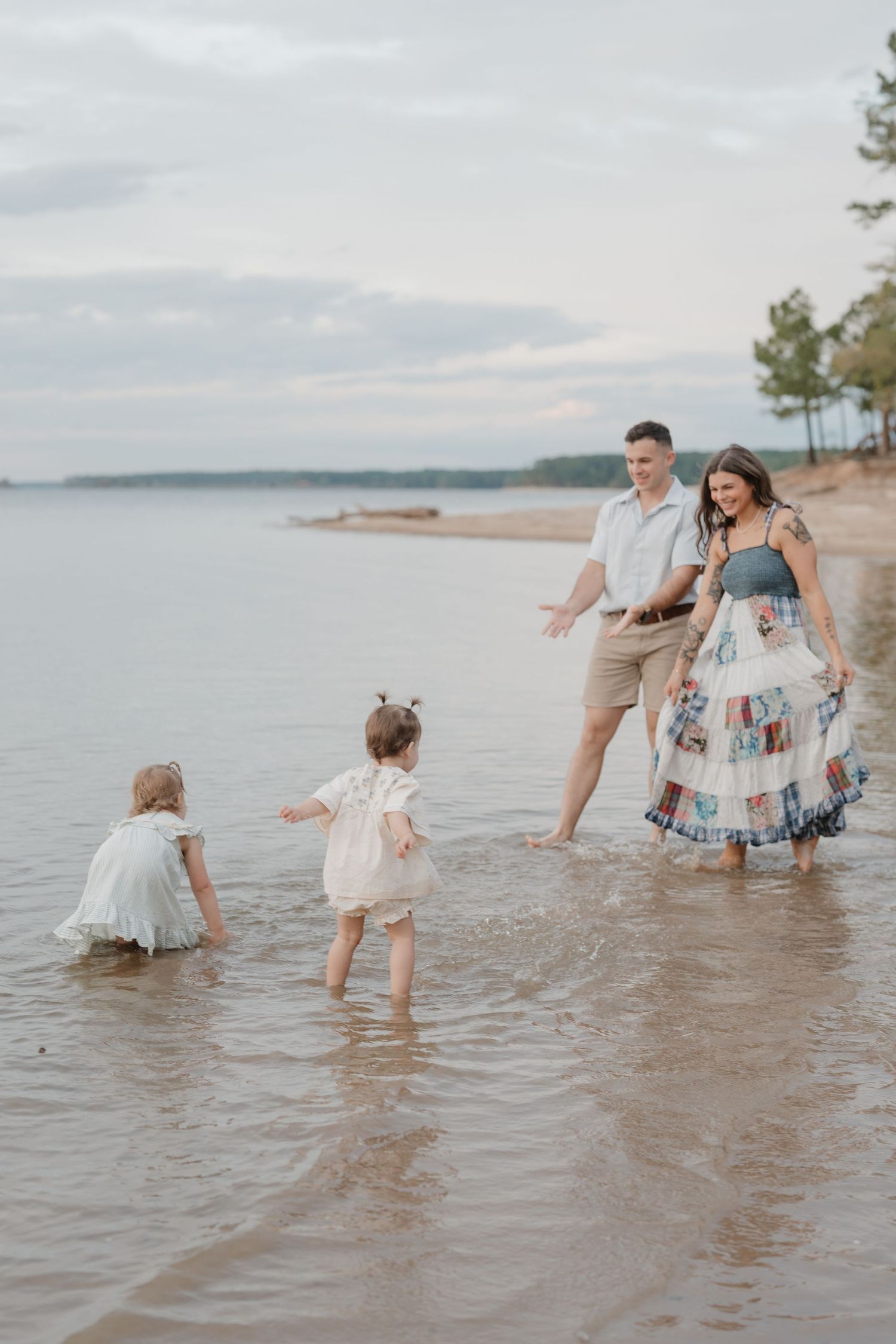 A family plays and splashes together in the shallow waters of a serene beach during a cloudy day.