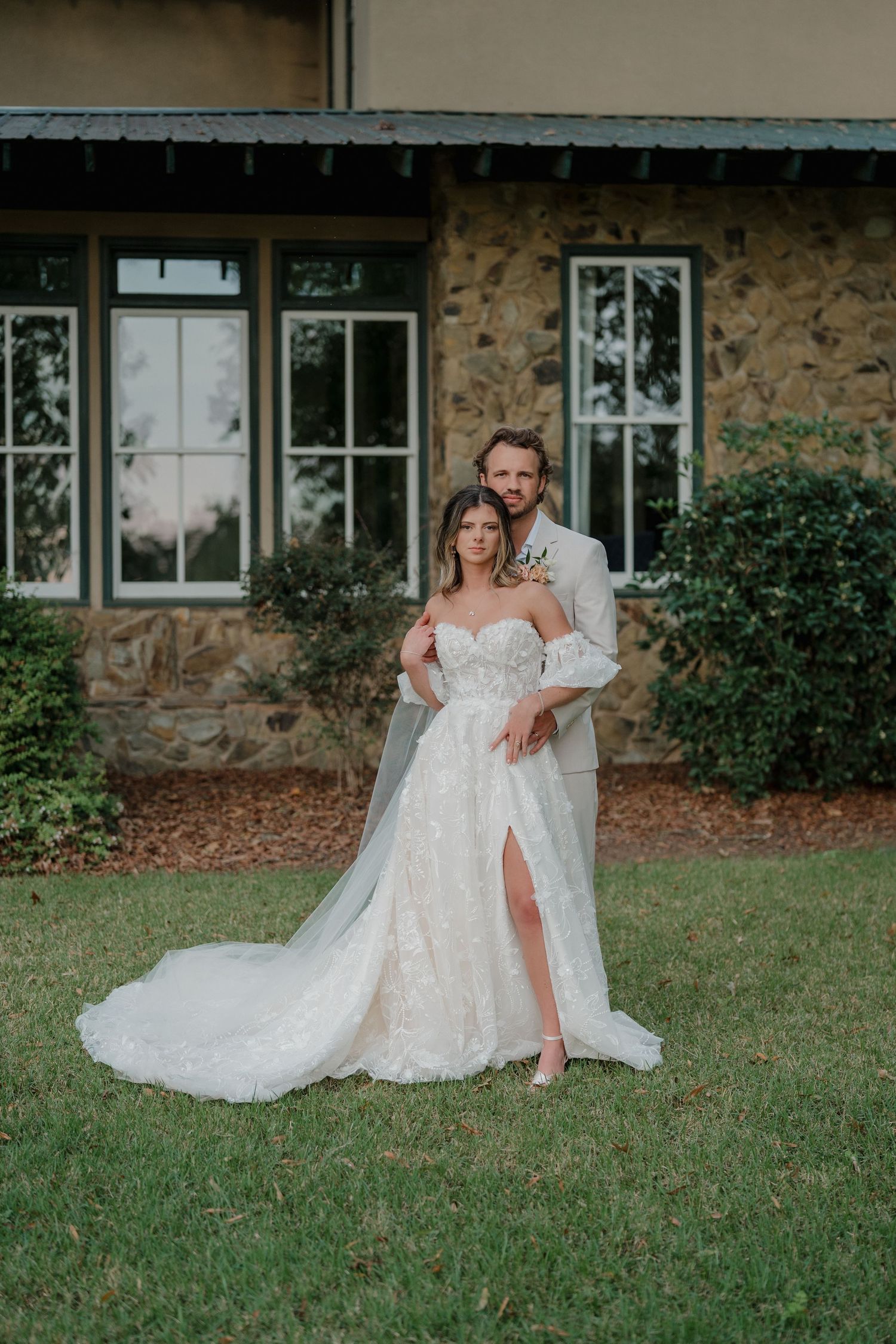 Elegant wedding couple poses in front of stone building with bride wearing lace gown with leg slit.