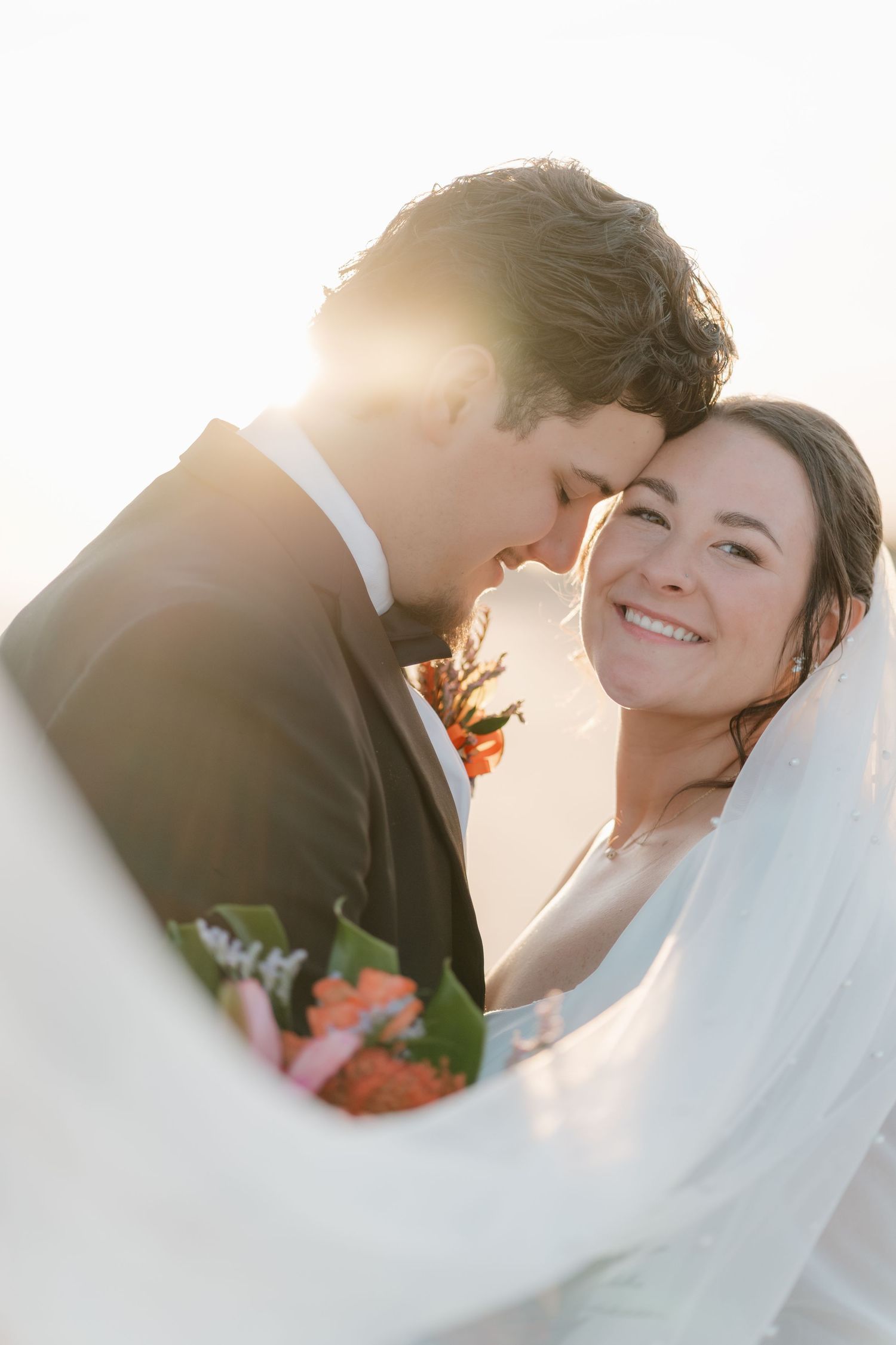 Intimate wedding portrait of newlyweds sharing tender moment at sunset with bridal veil flowing in foreground.