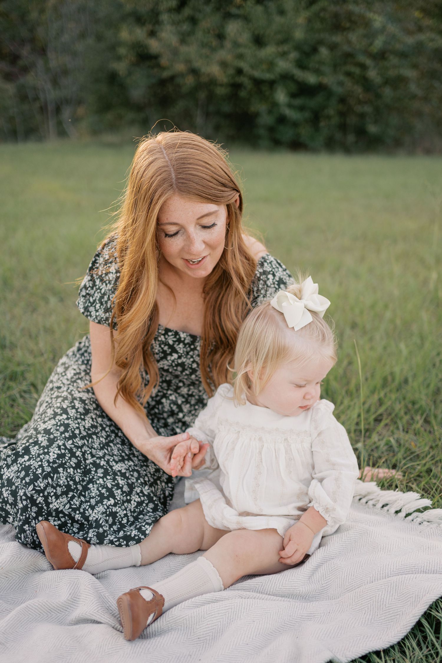 A mother and daughter share a tender moment sitting on a blanket outdoors, wearing floral and white dresses.