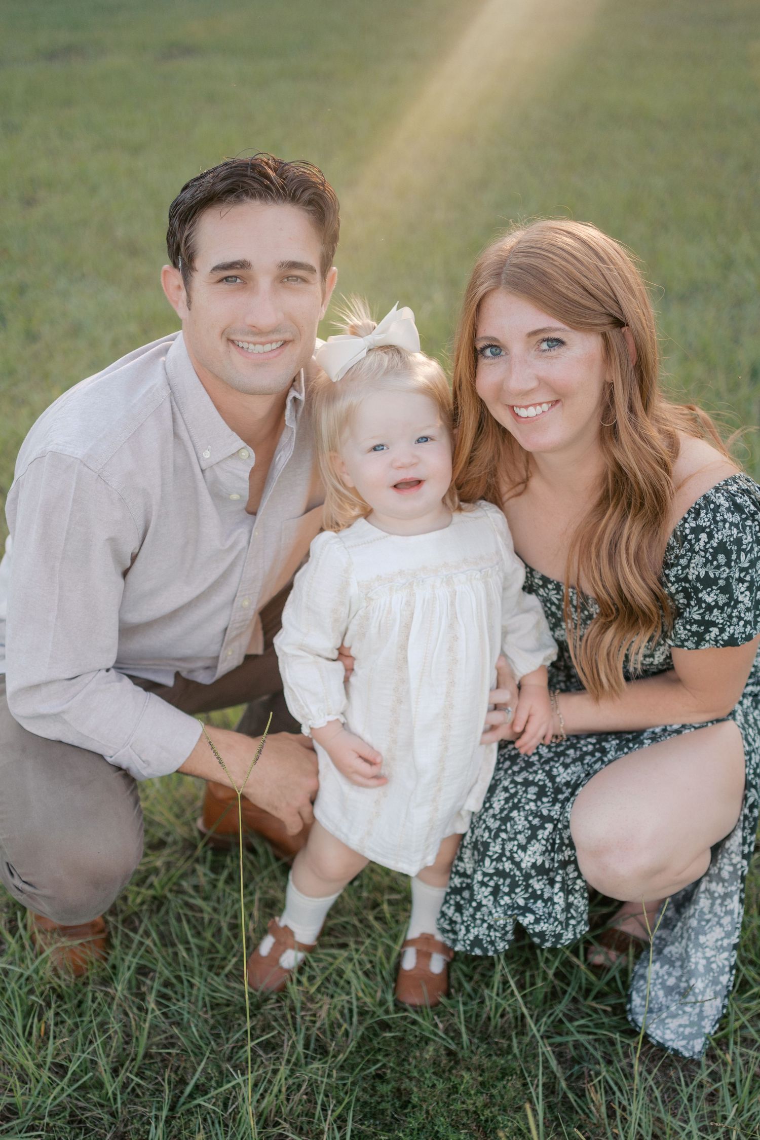 A family of three poses together on green grass wearing coordinated outfits in white and floral patterns.