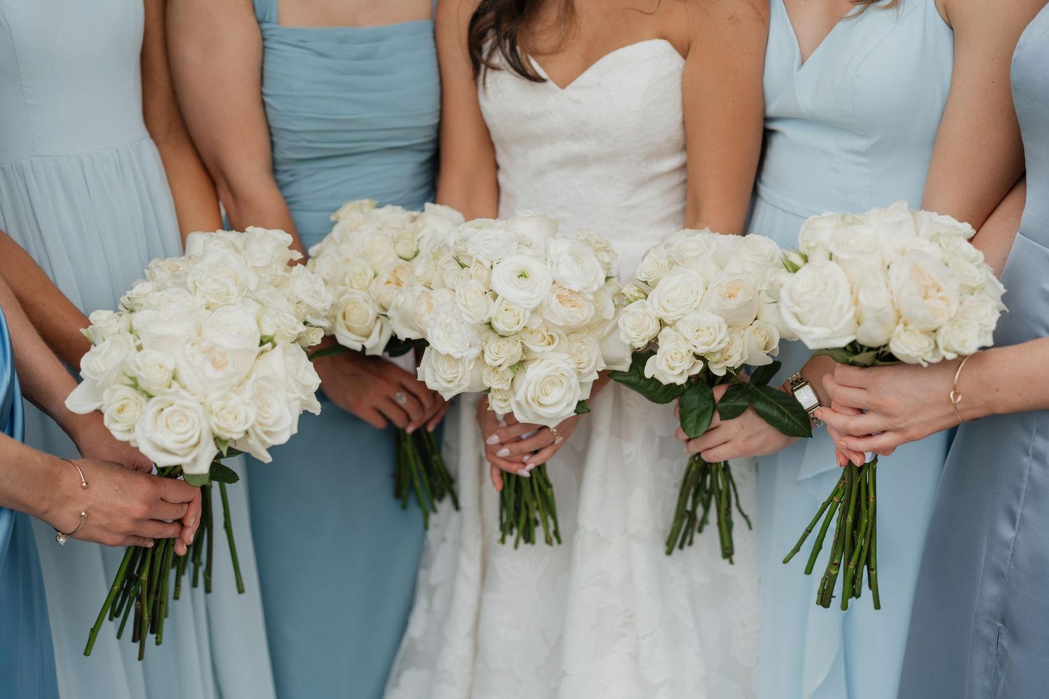 A row of bridesmaids in light blue dresses holding white rose bouquets stand together at a wedding.