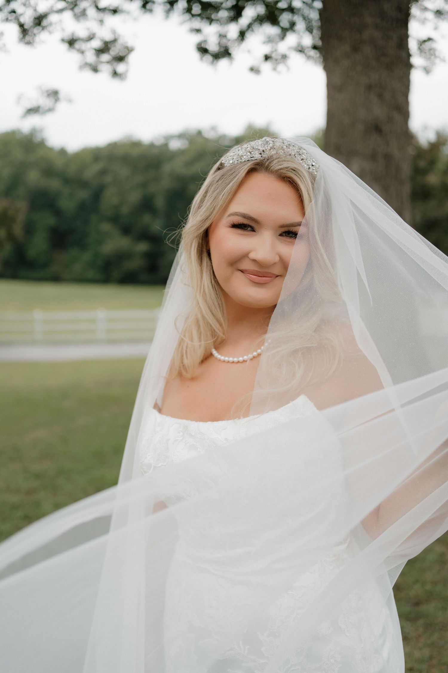 A bride in a white wedding dress and veil smiles outdoors with flowing sheer fabric caught in the breeze.
