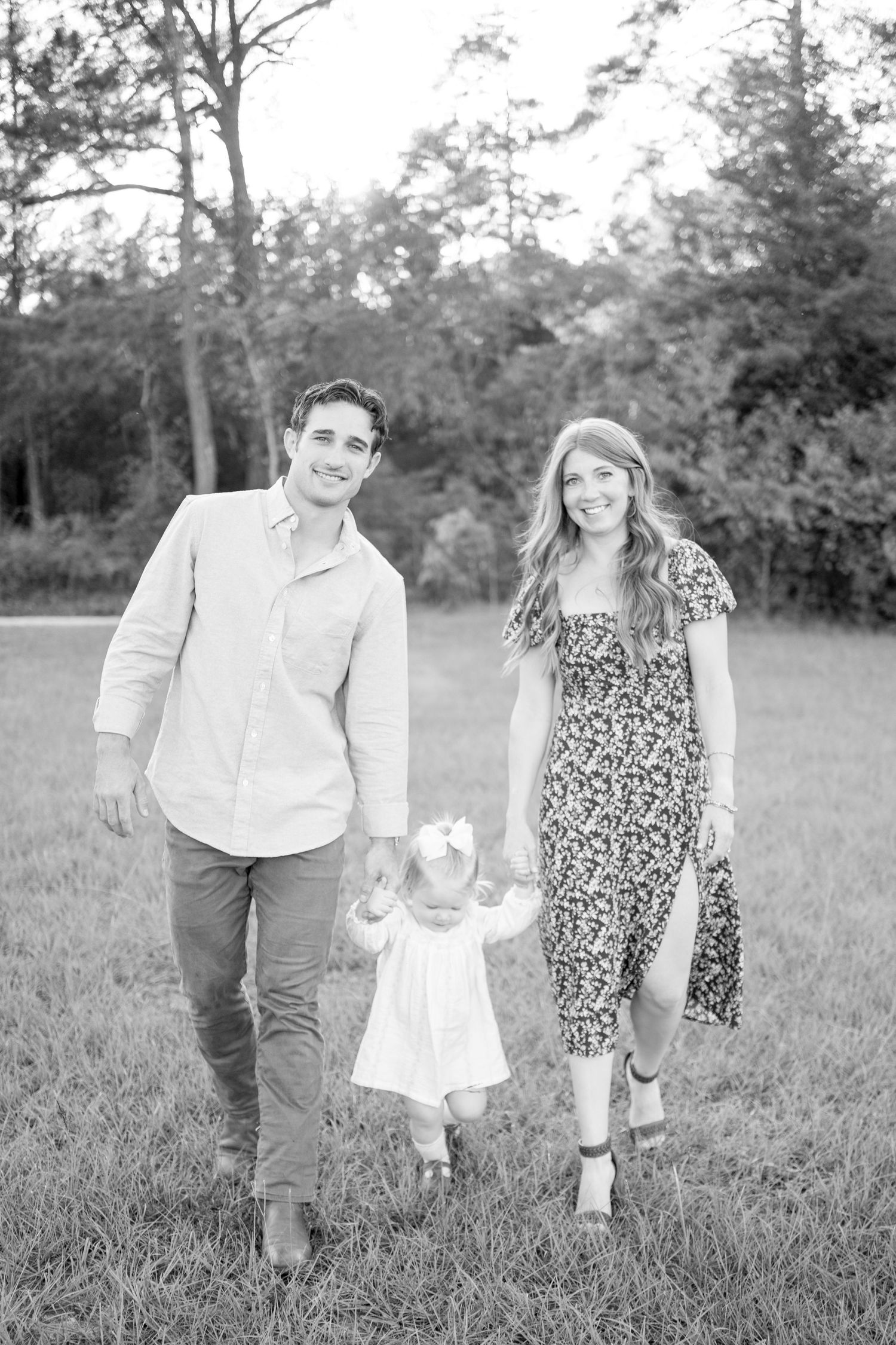Black and white outdoor family portrait showing parents walking hand-in-hand with a small child on a grassy field.