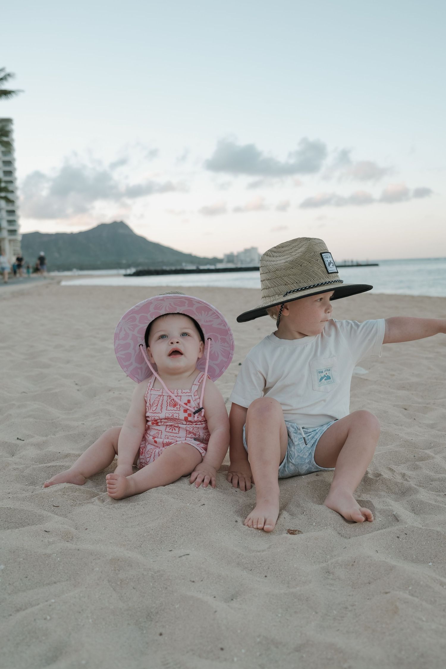 Two babies sitting on sandy Waikiki beach at sunset, wearing straw and pink sun hats.