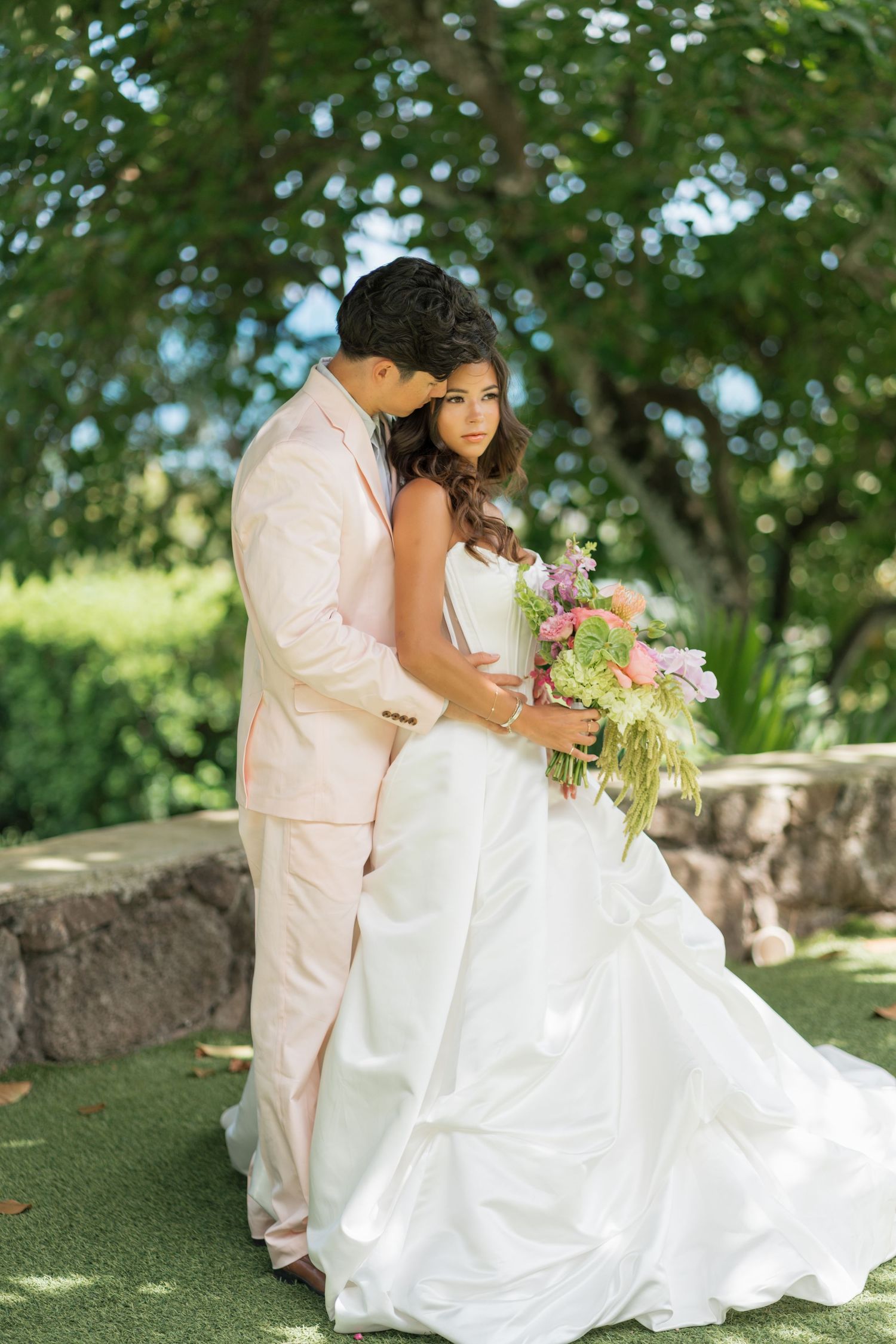 Bride in white gown holding pink bouquet stands with groom in blush suit outdoors under lush green trees.