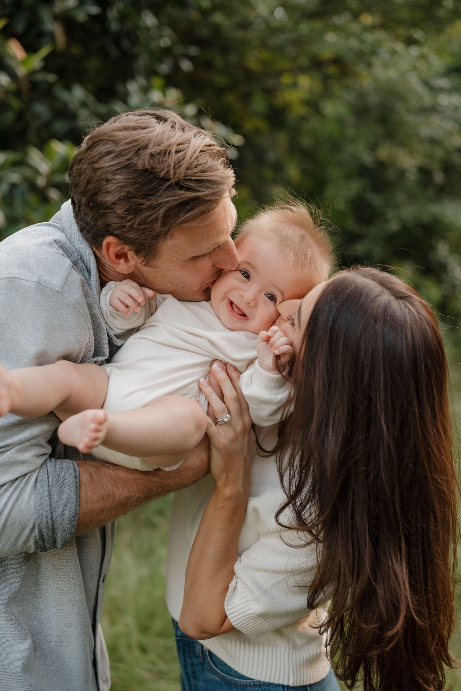 A loving family moment outdoors as parents share kisses and cuddles with their laughing baby in a natural setting.