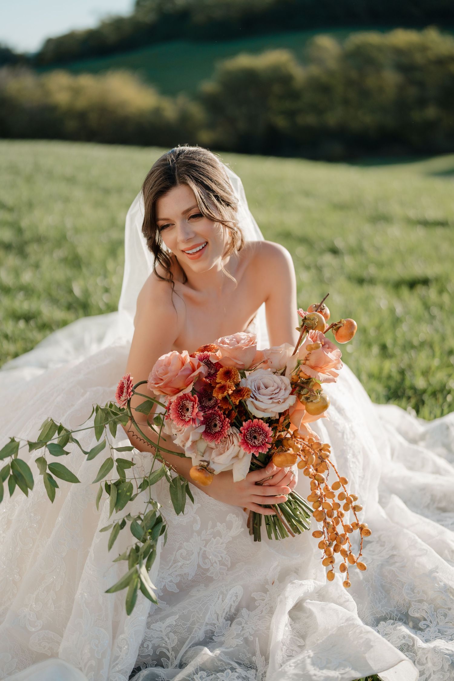 A bride in a white lace gown sits in a sunny meadow holding a vibrant coral and orange autumn wedding bouquet.