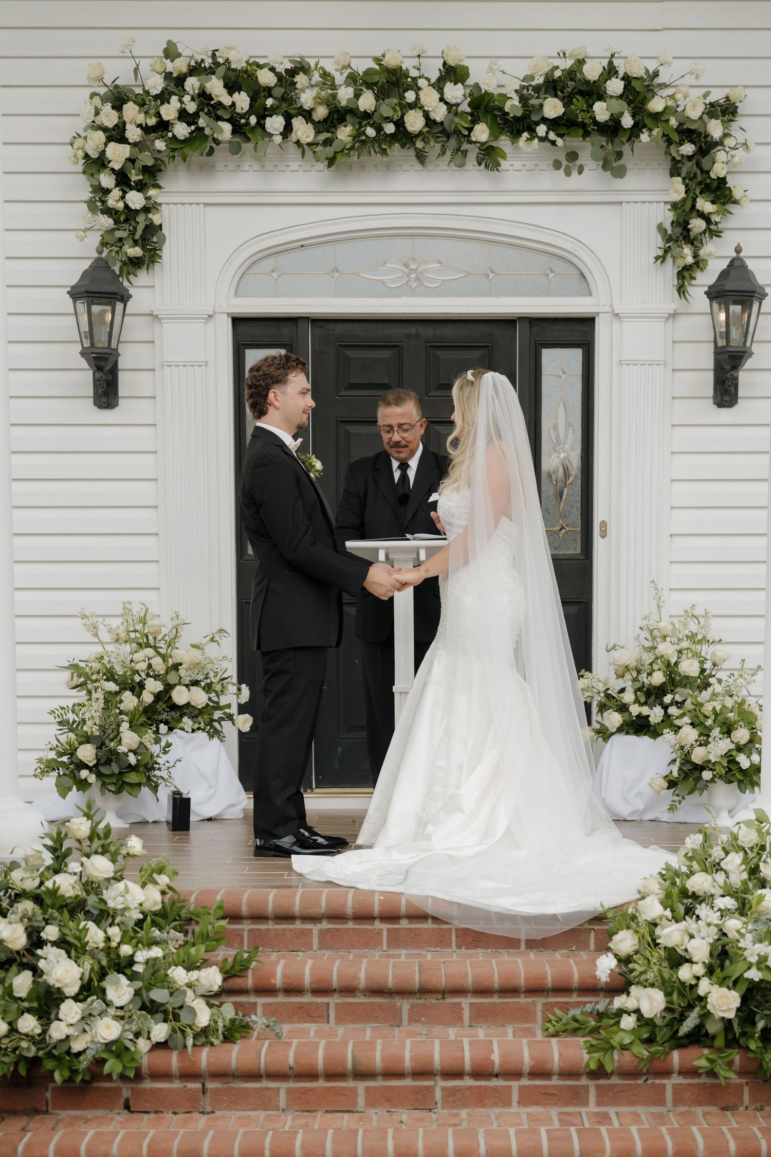 Wedding ceremony at a white church entrance with floral arch and brick steps as couple exchanges rings before officiant.