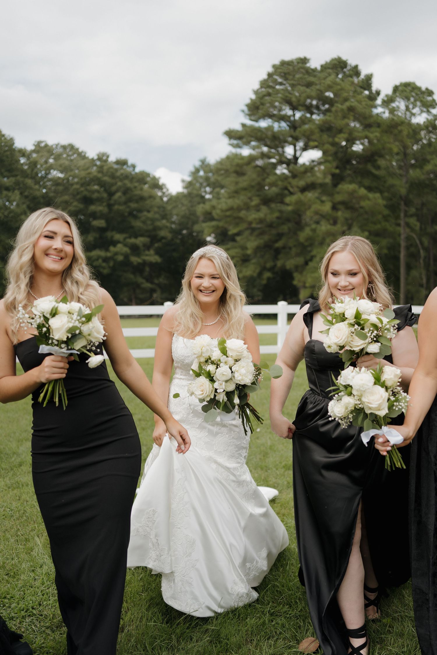 Wedding party members in black dresses carrying white bouquets walk alongside someone in a white gown on grass.