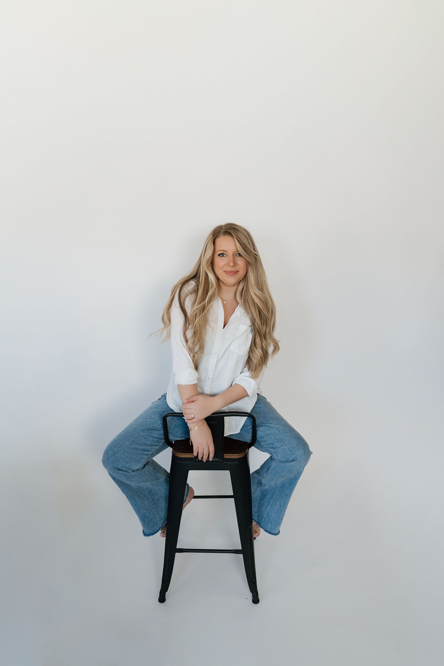 Person in white sweater and blue jeans sits casually on black stool against white background in studio setting.