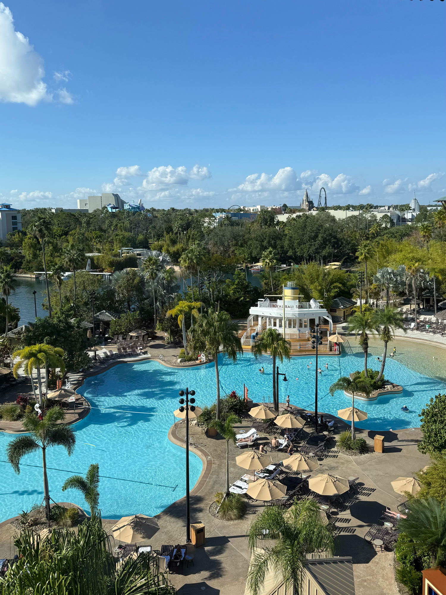 Aerial view of a resort pool with palm trees, lounge chairs, and umbrellas near a theme park in Florida.