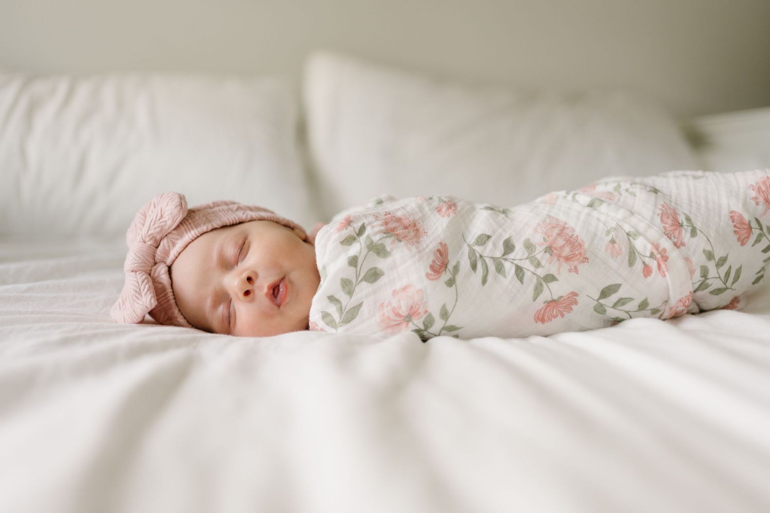 A sleeping newborn baby lies peacefully on white bedding with soft pink floral accents.