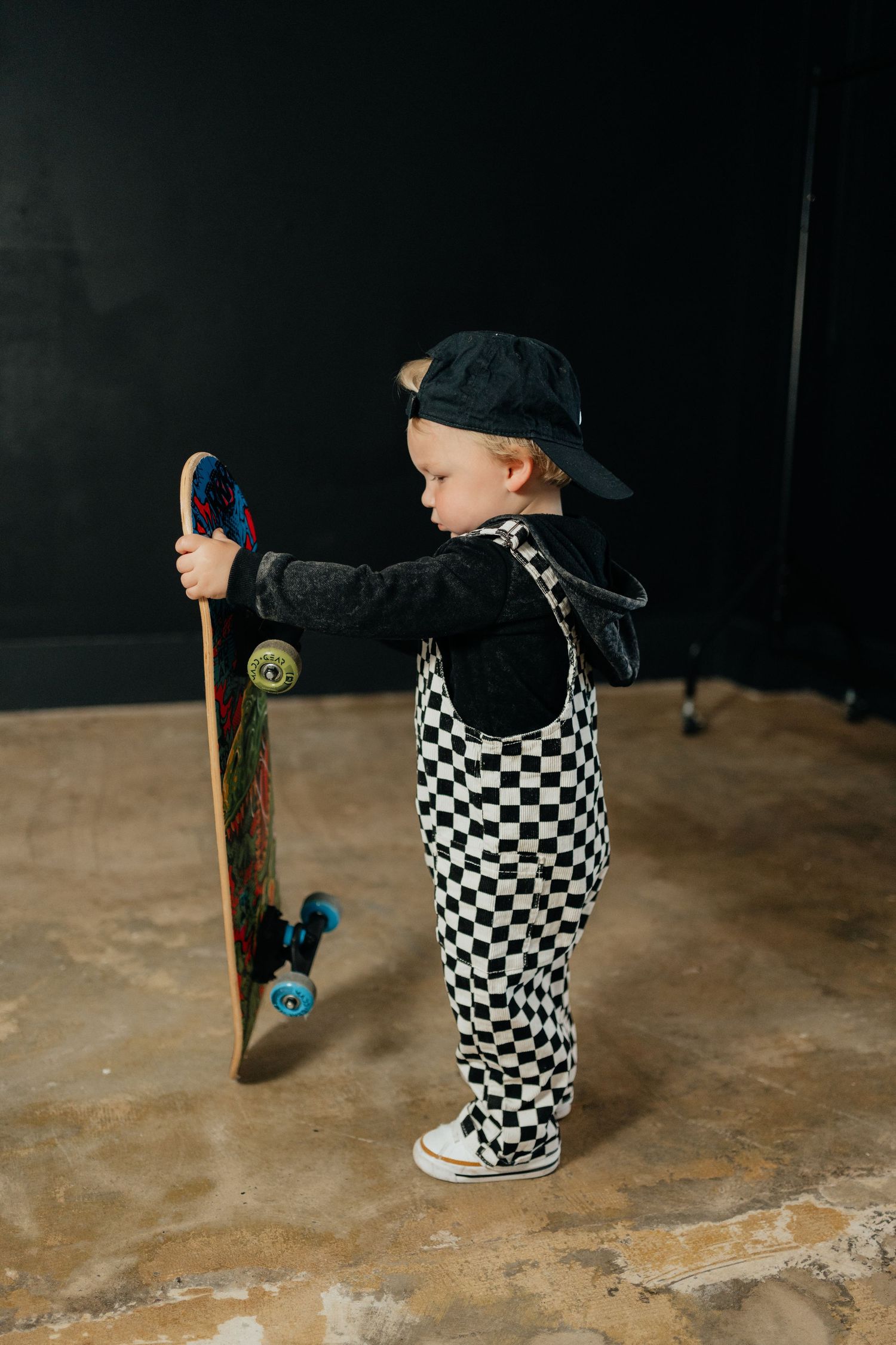 Toddler in checkered overalls and black cap holding a colorful skateboard upright on a studio floor.