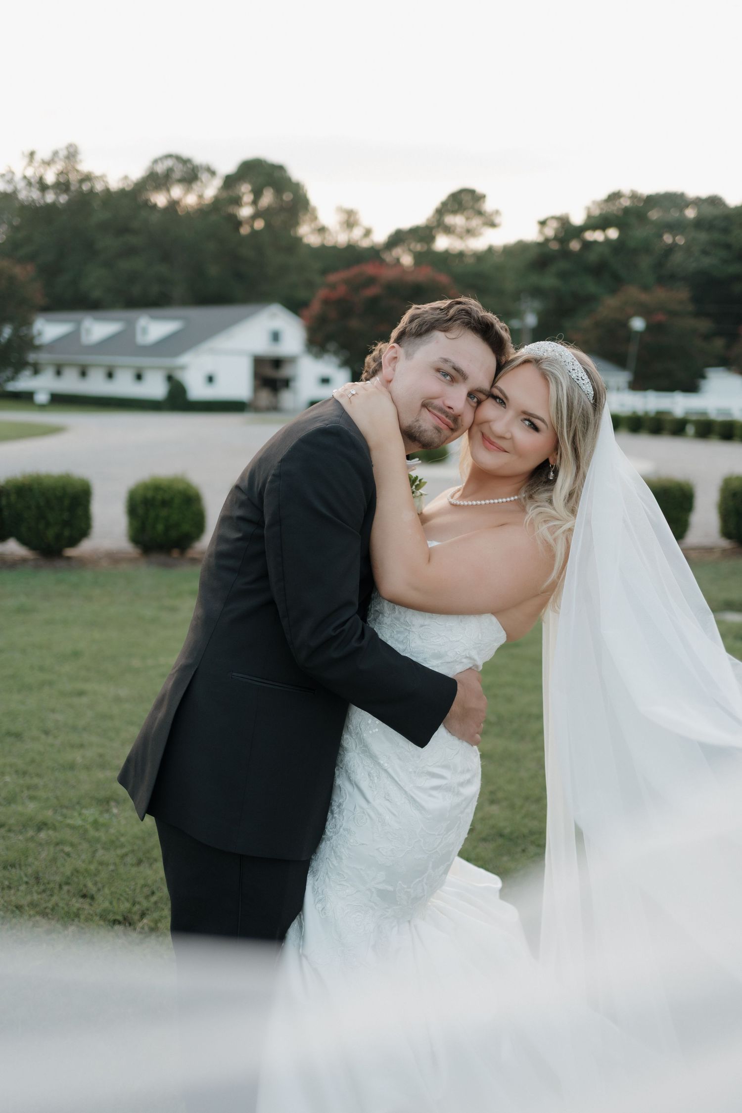 A happy couple embraces on their wedding day at a countryside venue with white buildings and trees in the background.