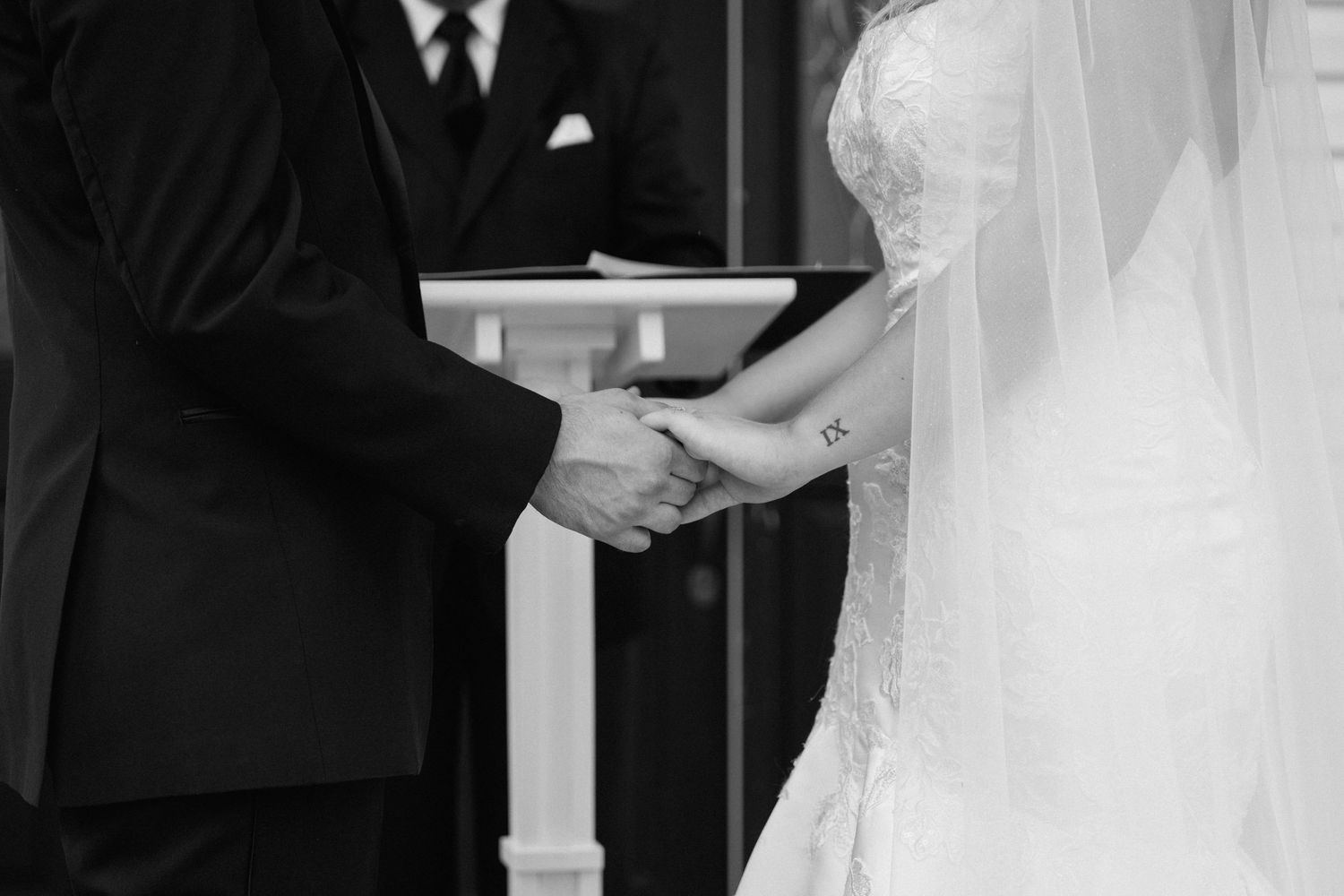 A black and white close-up photo shows hands clasped together during a wedding ceremony in front of a white pillar.