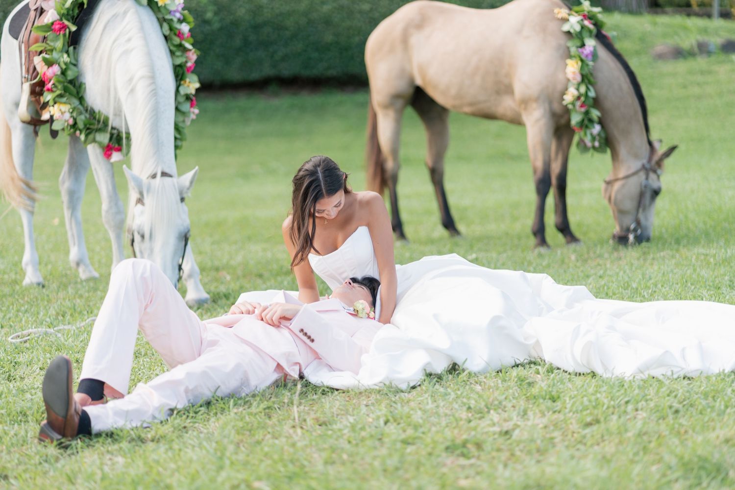 Bride and groom relaxing on green grass with decorated horses grazing nearby at an elegant outdoor wedding celebration.