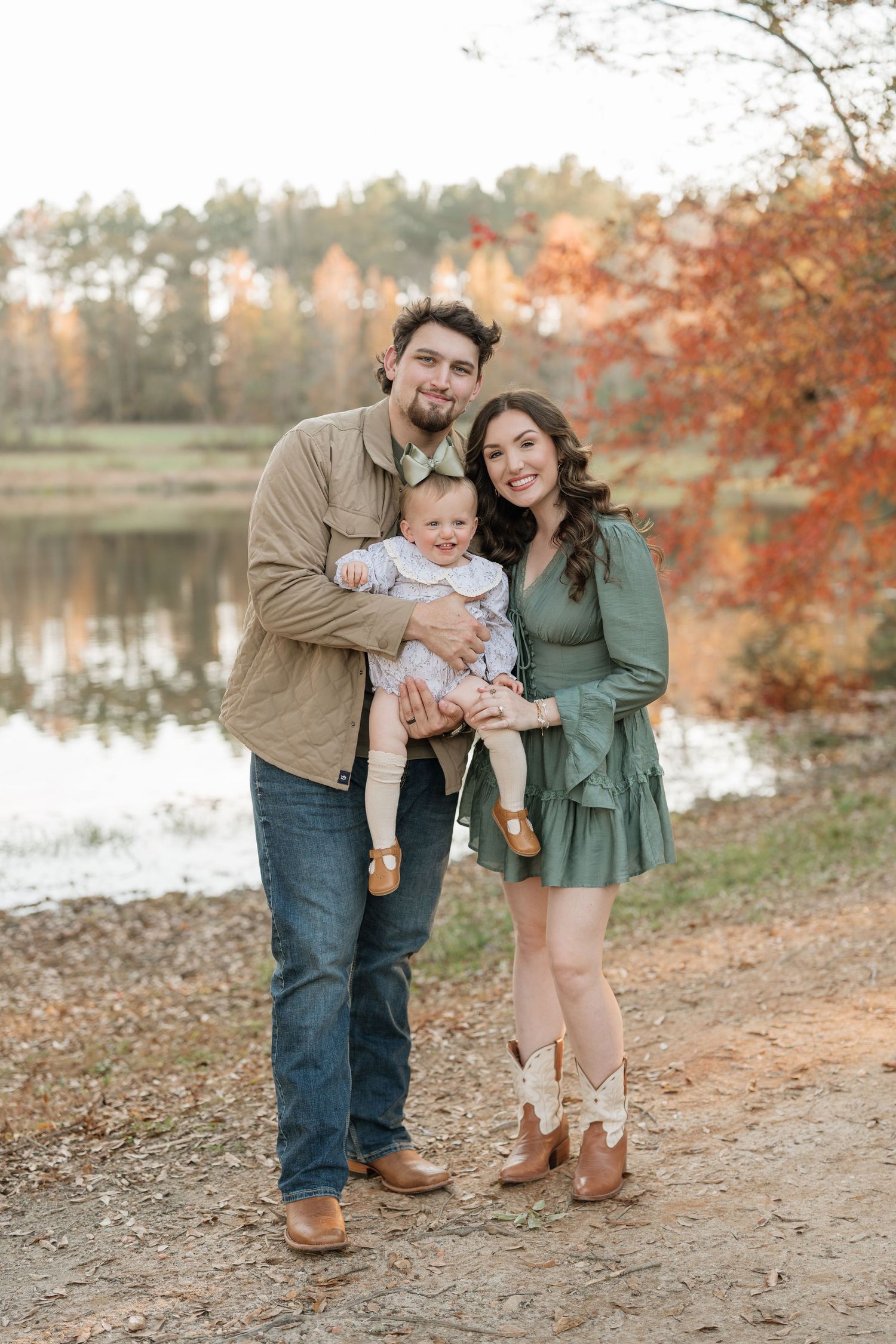 A family enjoys an autumn photoshoot by a lake with fall foliage and warm earth tones in their coordinated outfits.