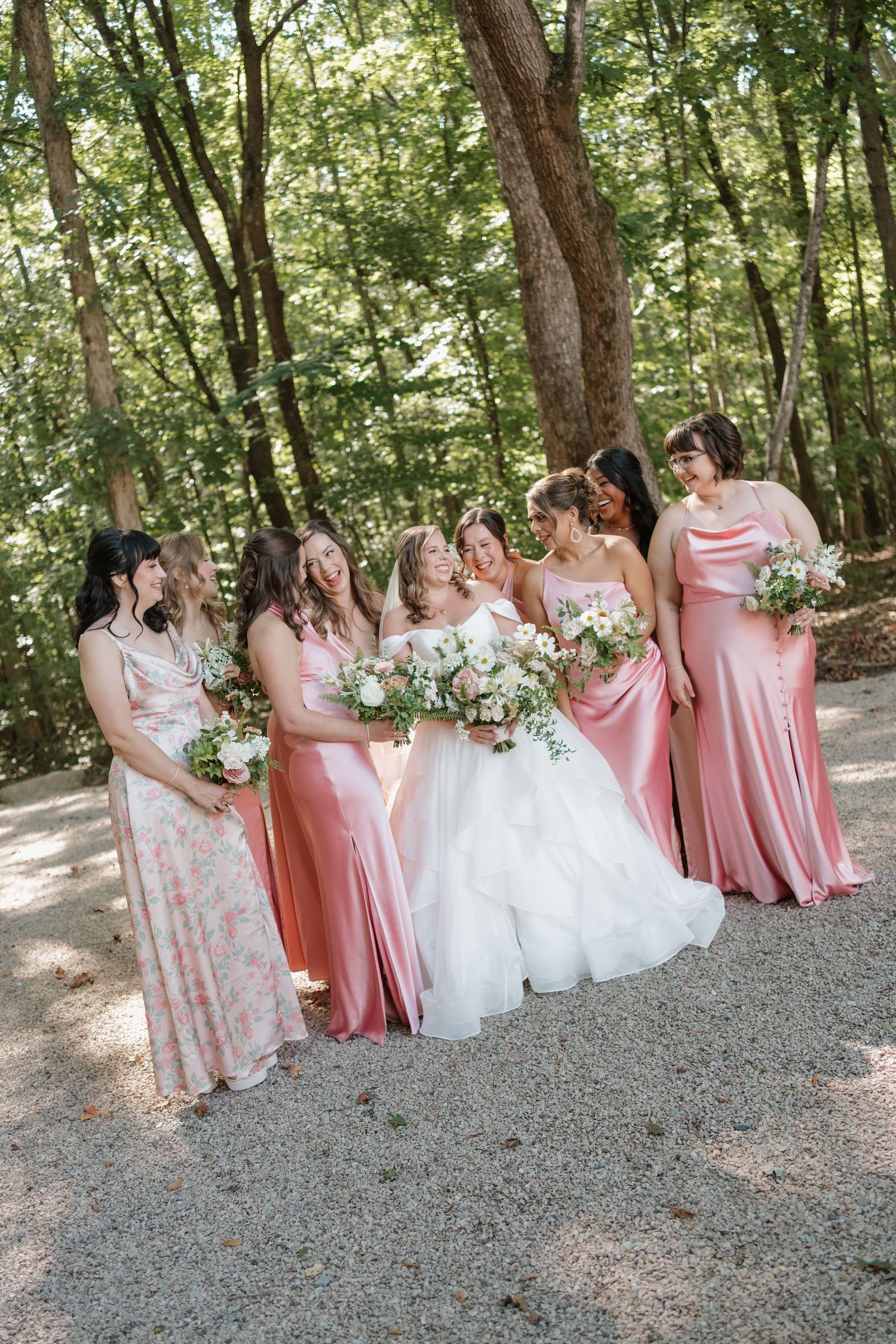 A bridal party in pink dresses and a white wedding gown pose together on a gravel path surrounded by trees.