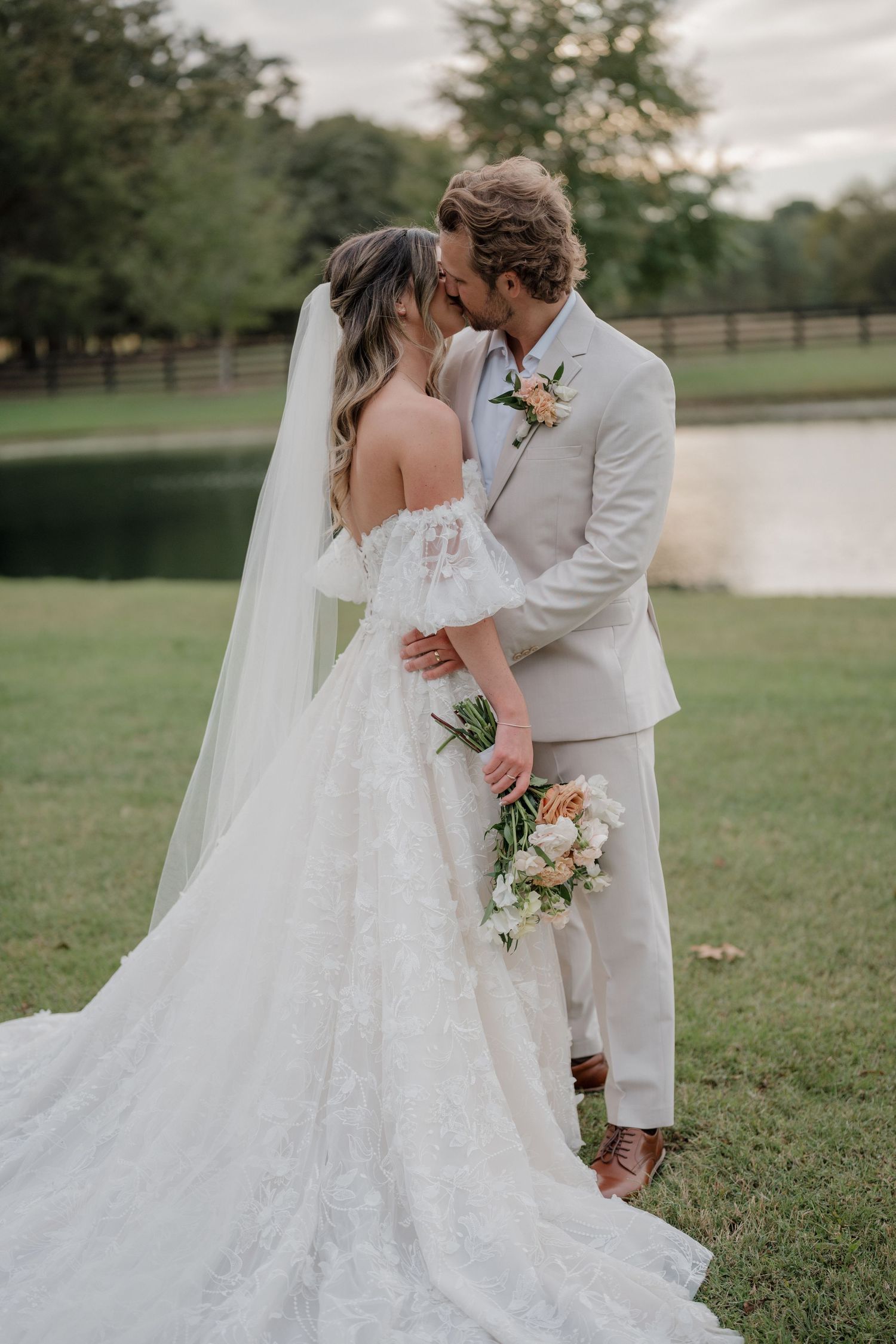 Elegant wedding couple poses in a grassy field near a lake with the bride's dress trailing behind.
