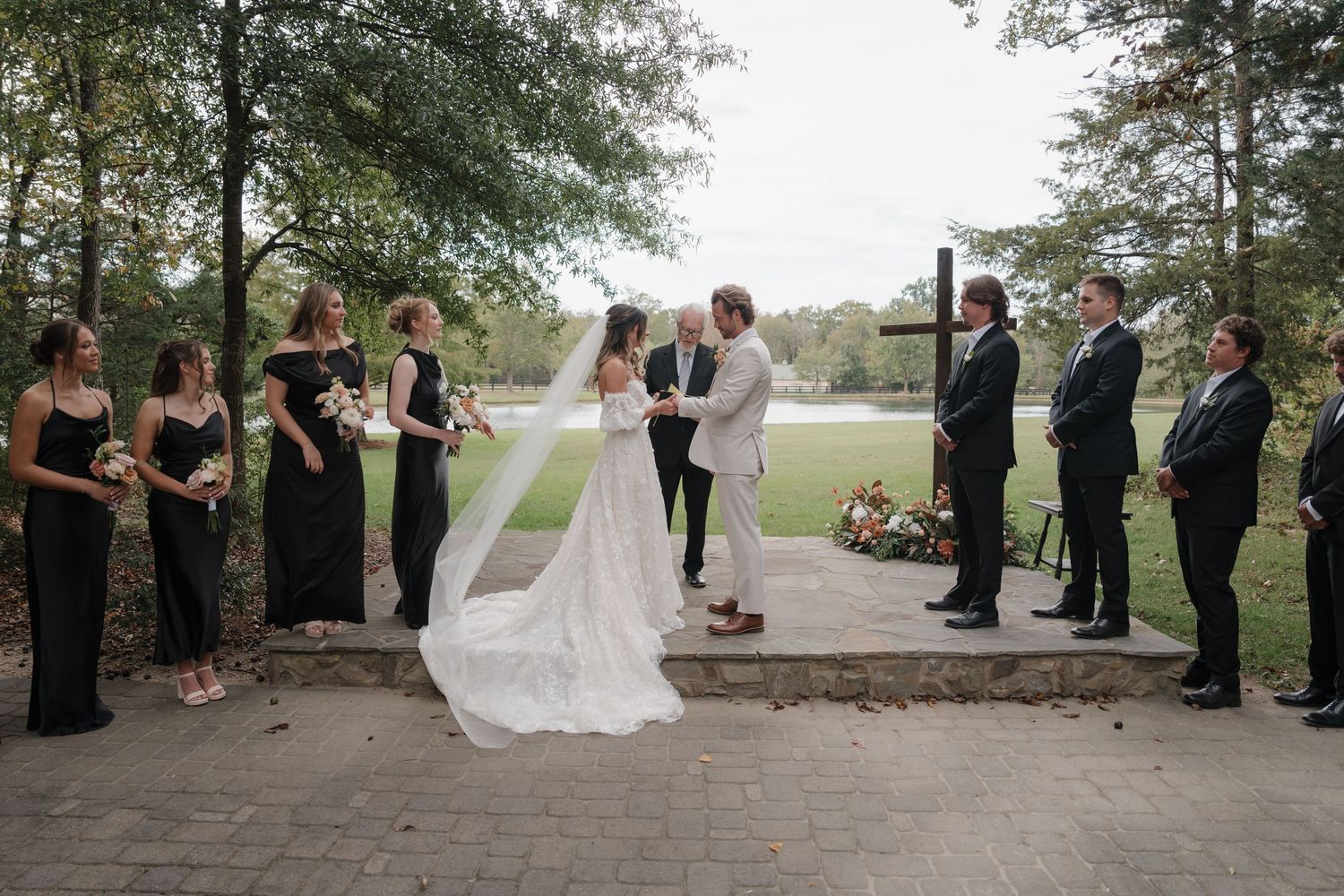 A wedding party stands in a line outdoors under tall trees during a formal ceremony.