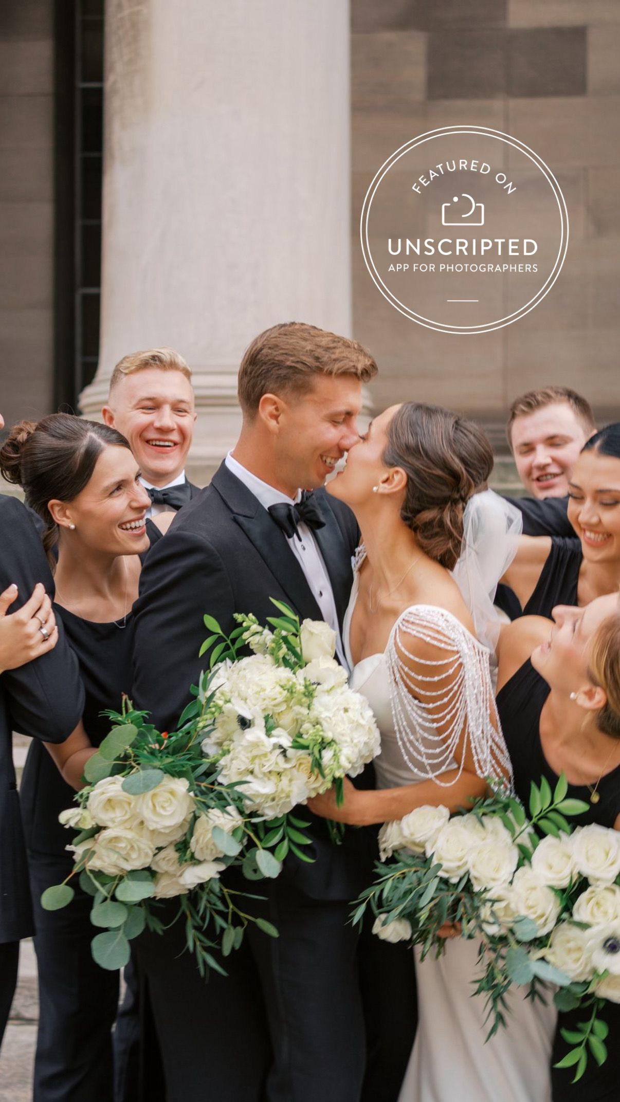 Wedding party laughs together while holding elegant white floral bouquets with eucalyptus against stone wall.
