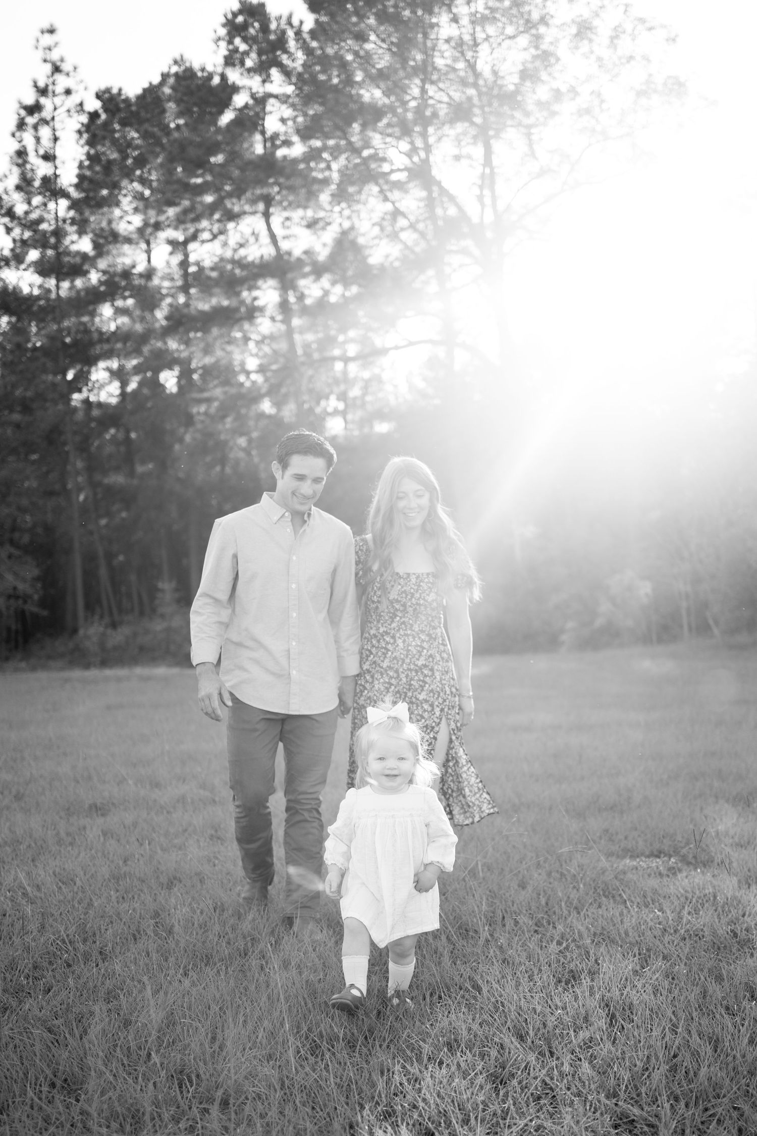 A family of three walks together in a sunlit grass field with trees in the background, captured in black and white.