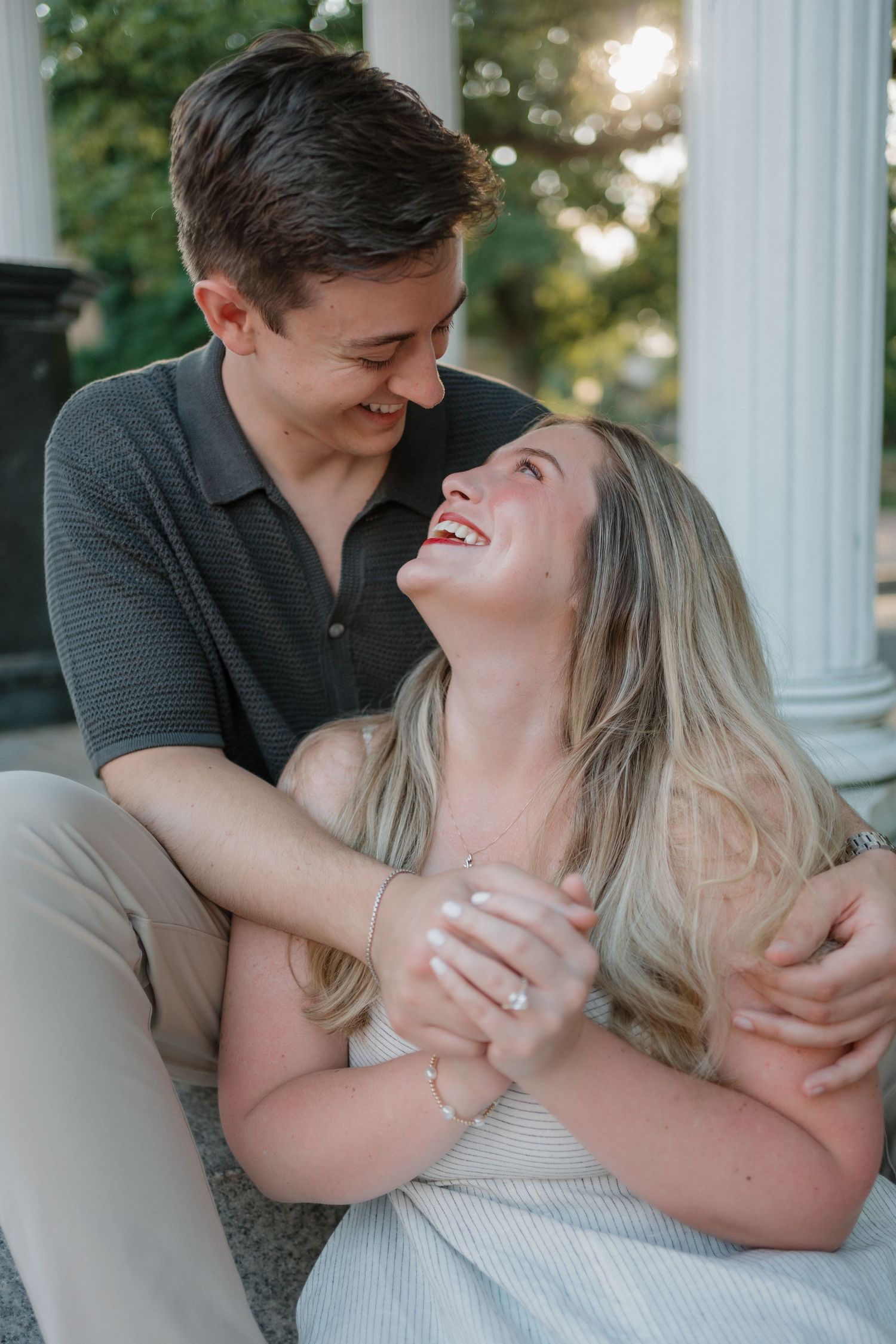 A joyful couple shares a candid laughing moment together while sitting outdoors during a sunny engagement photoshoot.