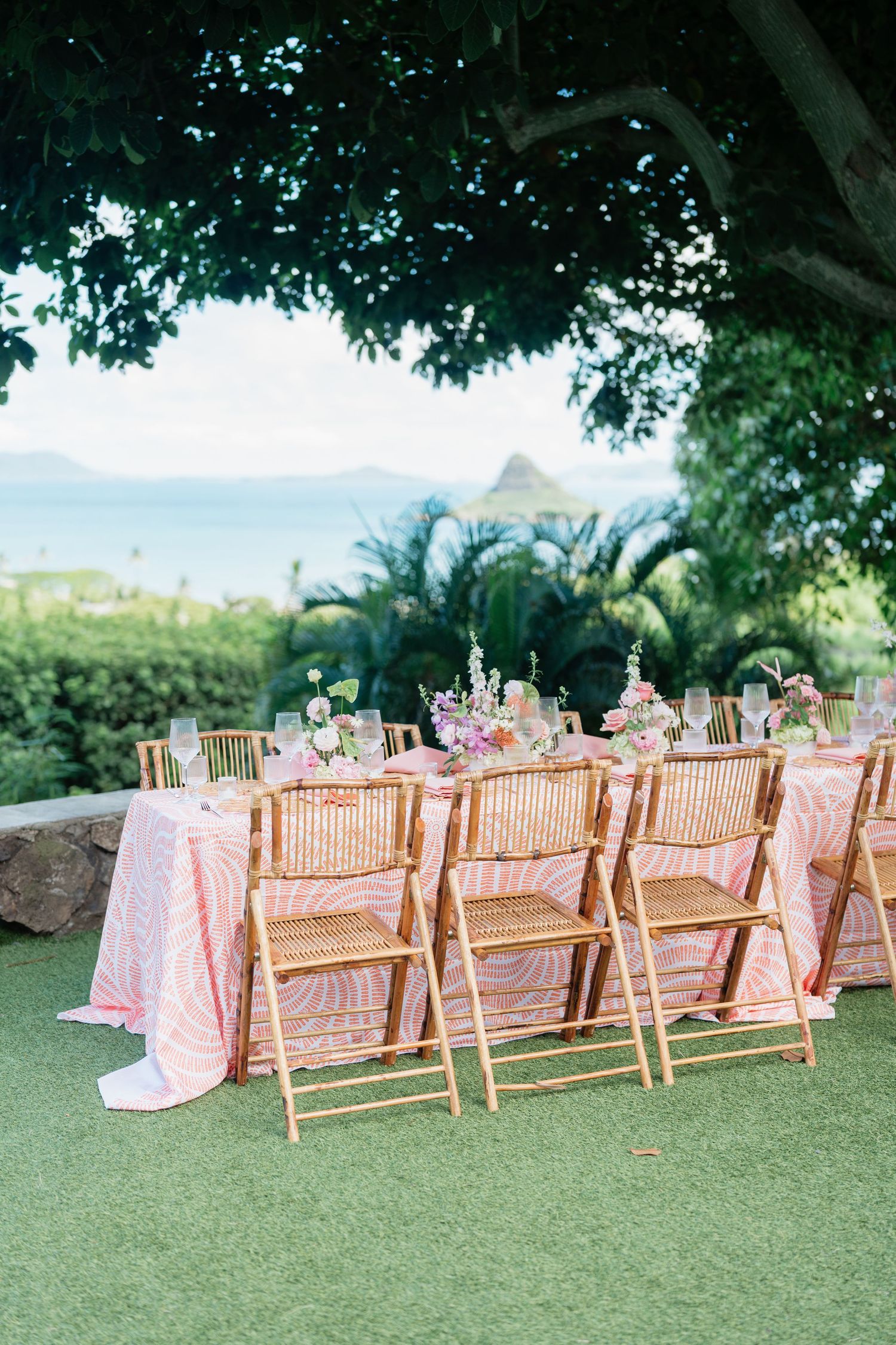 Outdoor wedding reception with bamboo chairs and pink tablecloths set on green lawn with tropical ocean views.