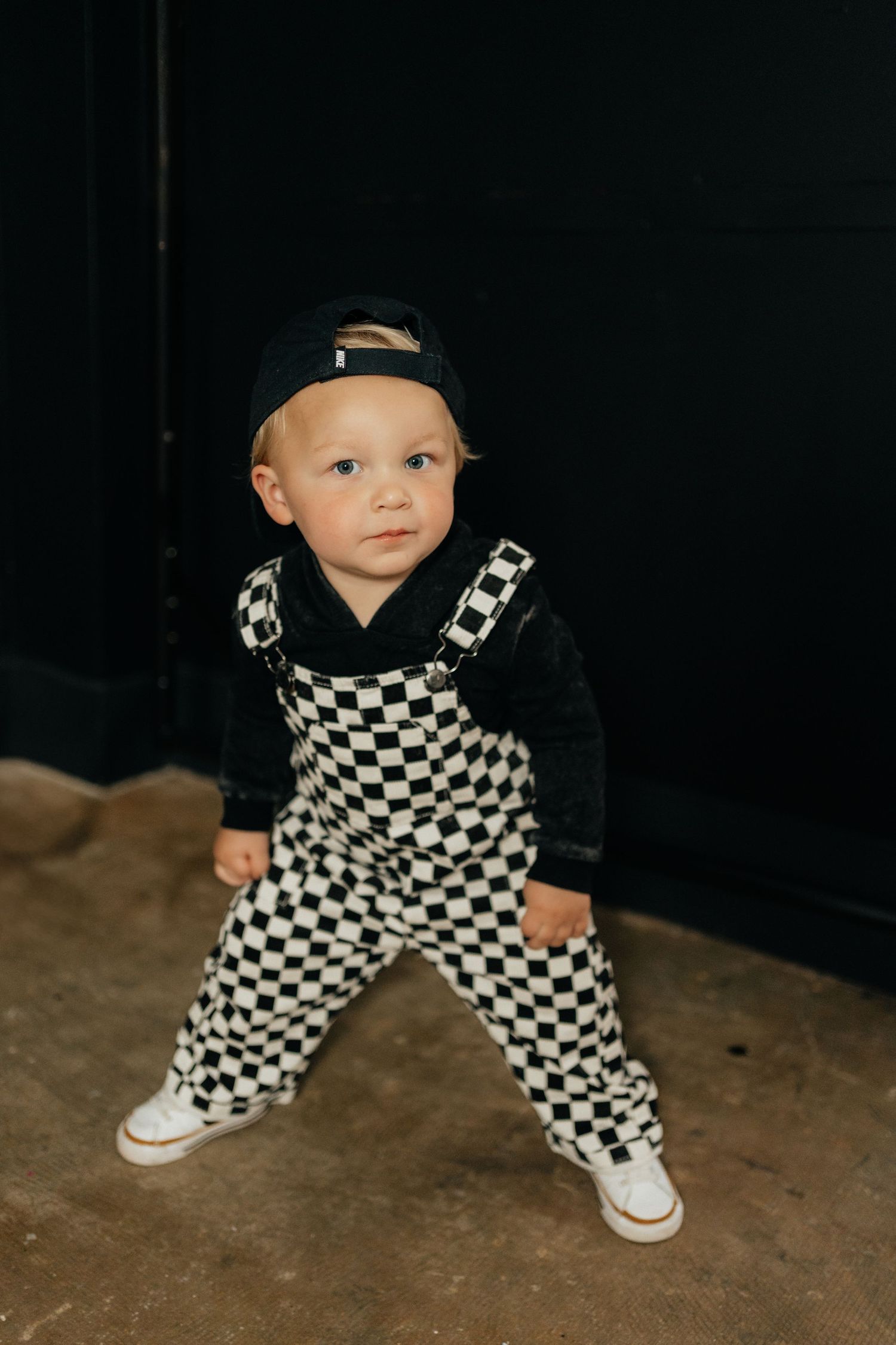 Toddler in black and white checkered overalls, black hoodie, and cap standing confidently on a concrete floor.
