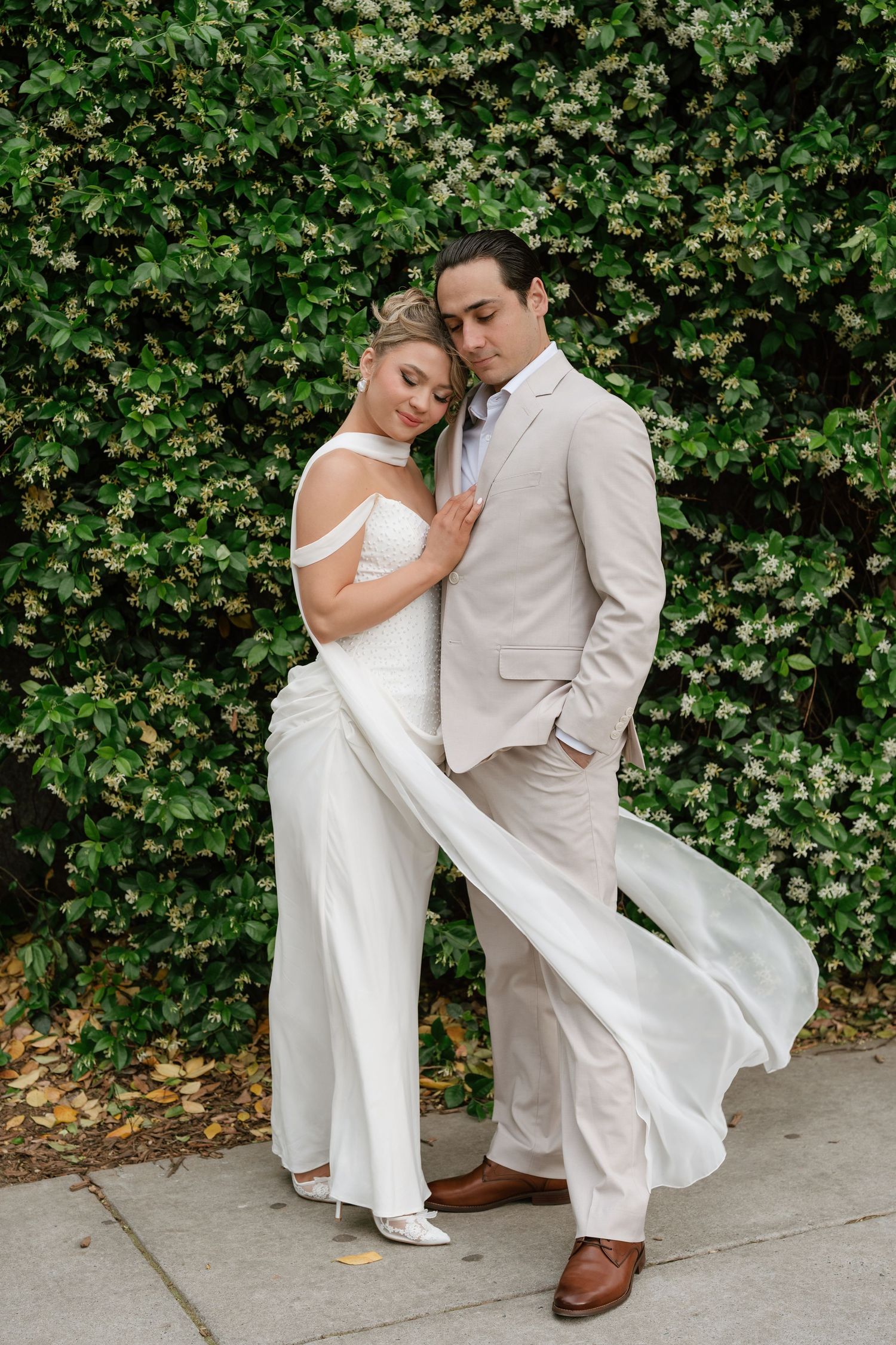 Bride in white off-shoulder gown embraces groom in beige suit before lush green floral wall.