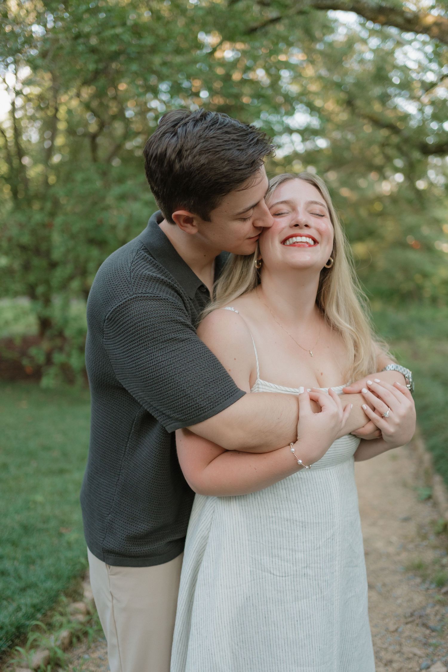 A romantic outdoor portrait of a couple embracing and laughing together on a nature path surrounded by green foliage.