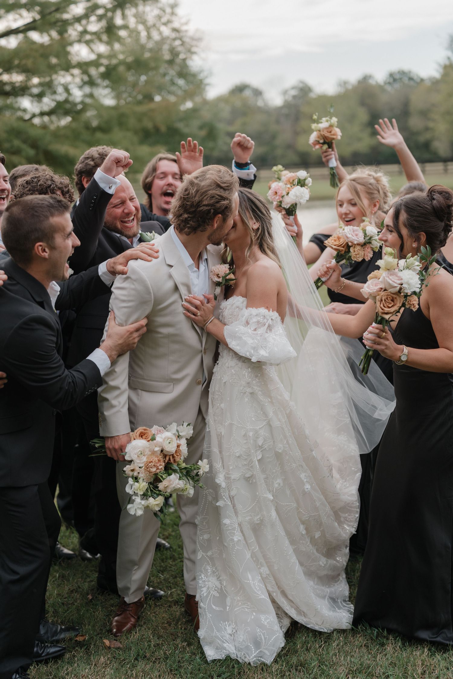 Wedding party celebrates with joyful cheers and raised hands as newlyweds walk through crowd during outdoor celebration.