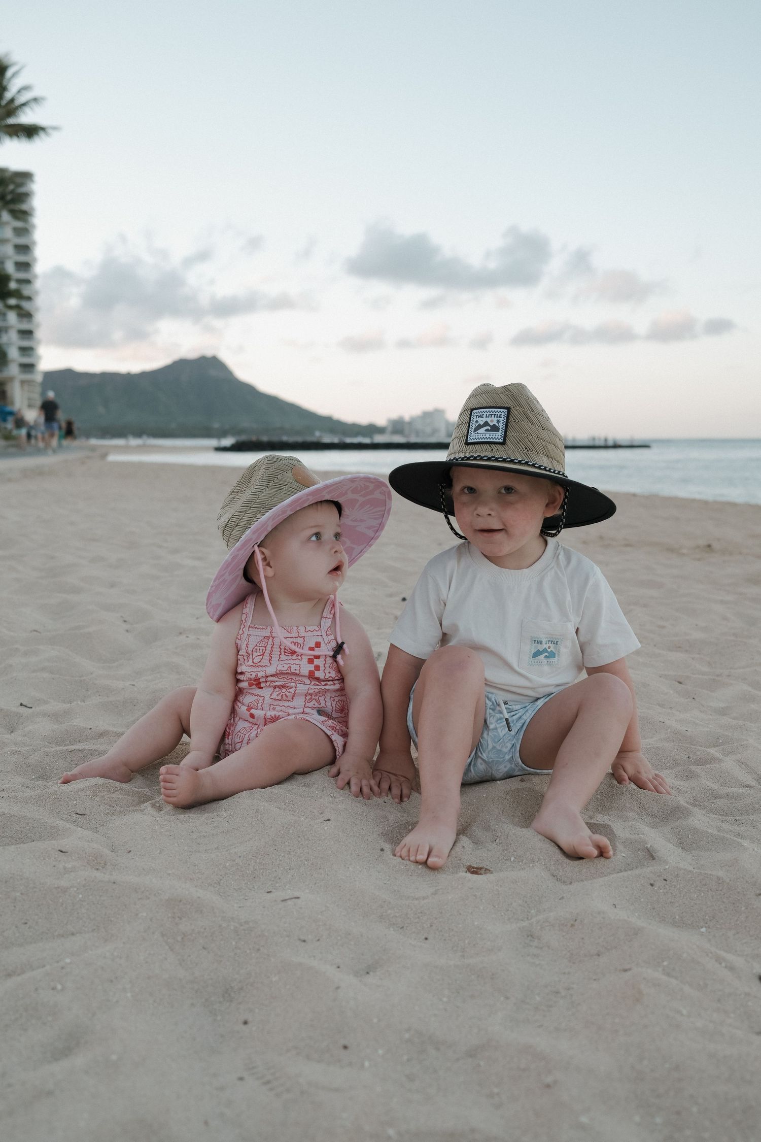 Two toddlers in wide-brim hats sit on Waikiki Beach sand with Diamond Head crater visible in the background.