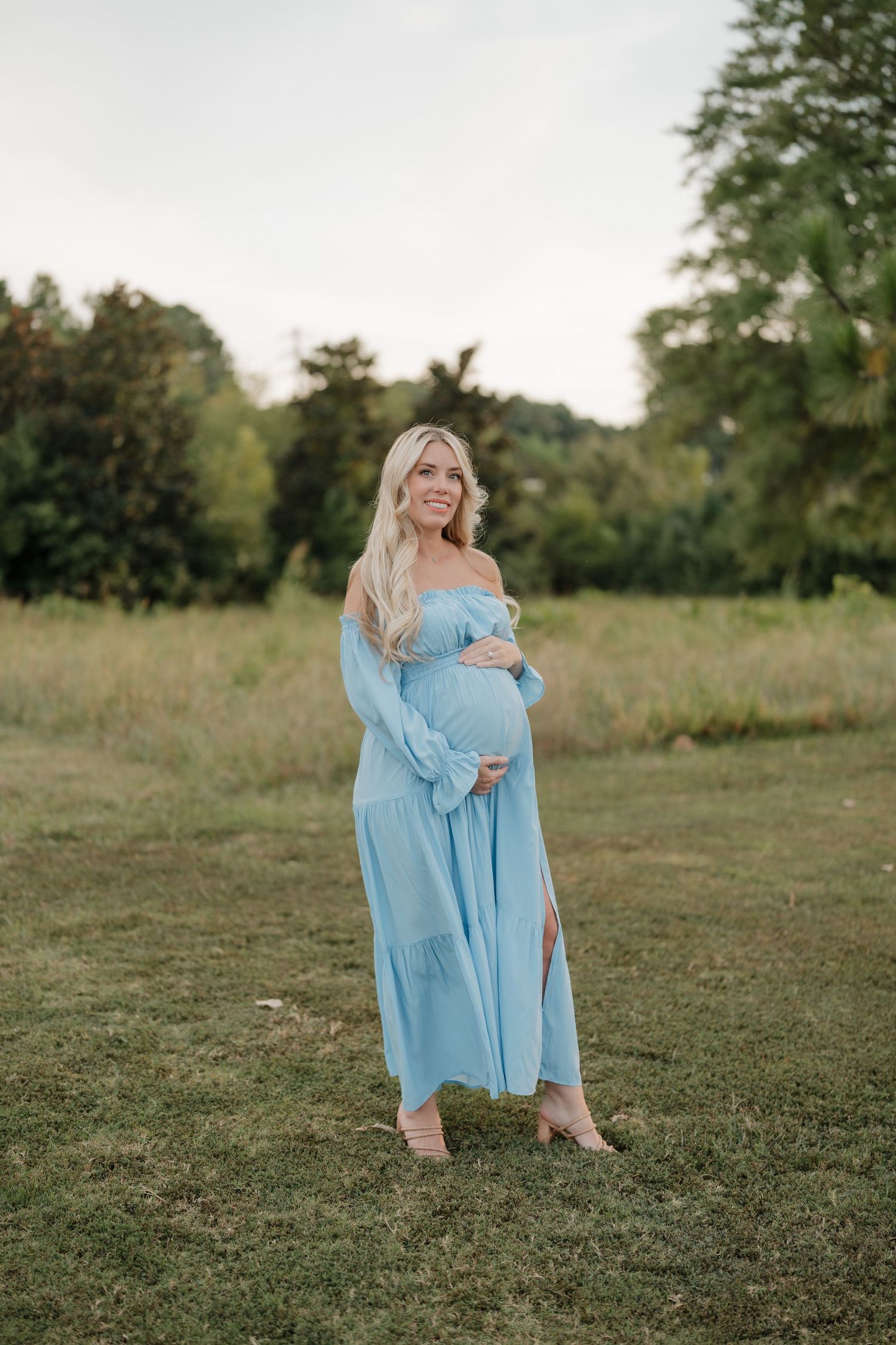 A mother and child in matching light blue dresses embrace while standing in a grassy meadow at sunset.