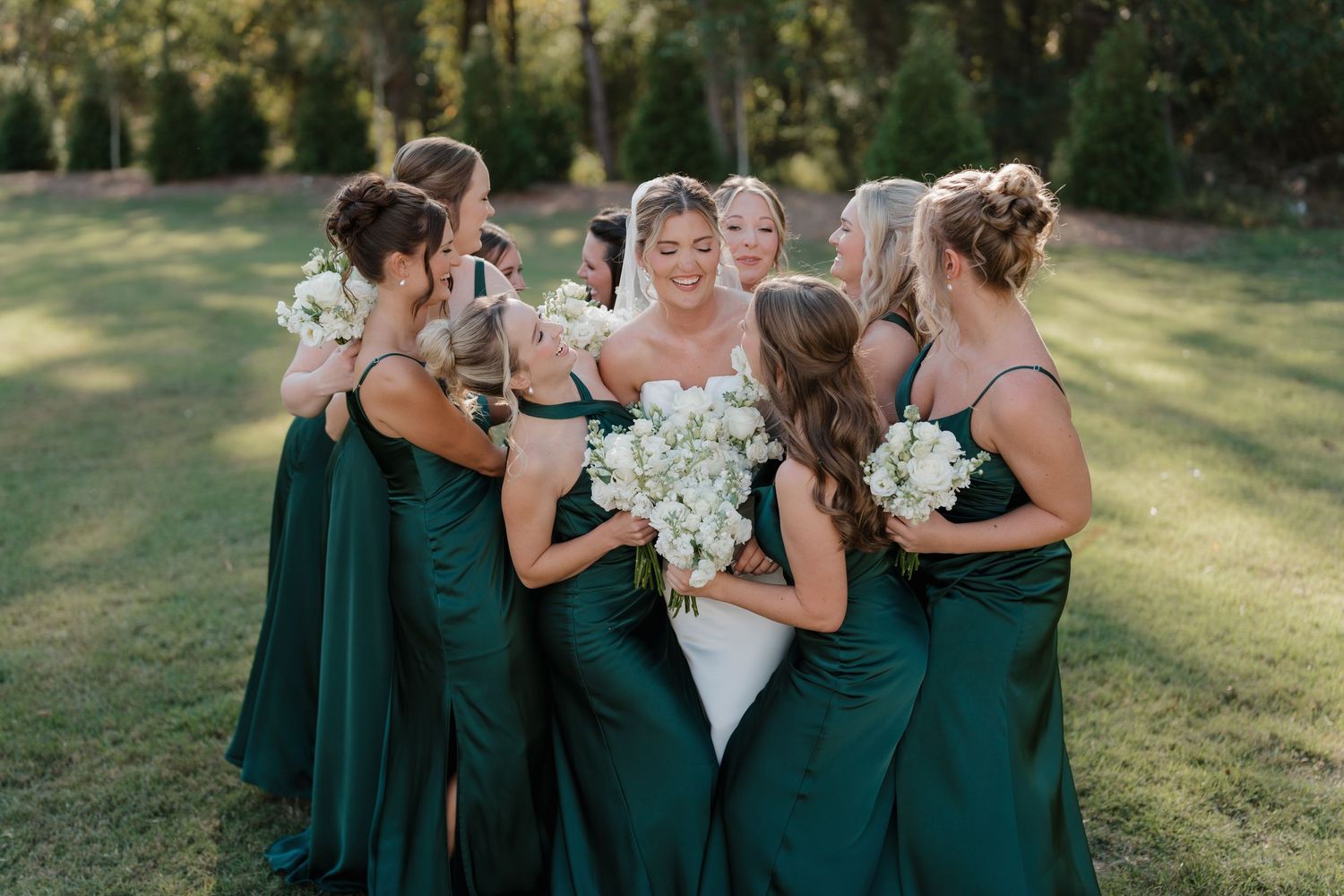 A group in emerald green dresses stands together holding white floral bouquets in a sunlit outdoor setting.