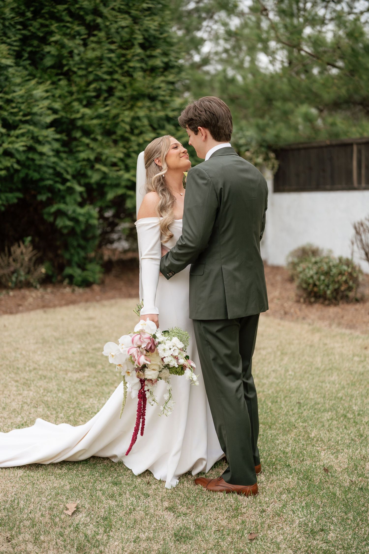 Bride in white off-shoulder gown holding floral bouquet embraces groom in olive suit outdoors on green lawn.