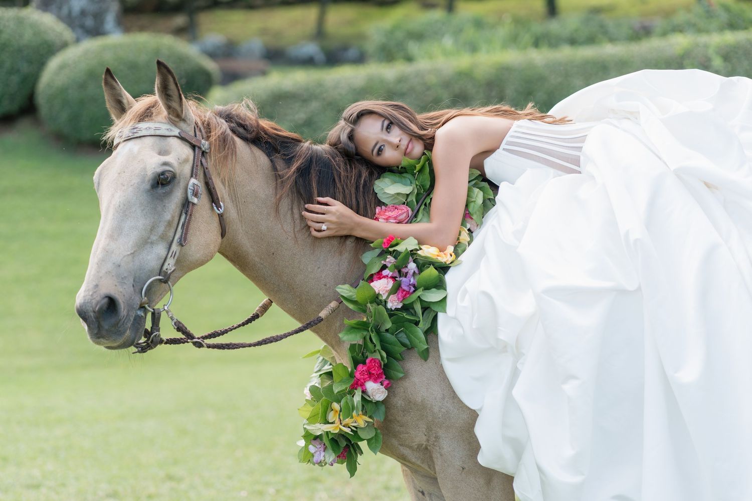 Bride in white gown drapes over a decorated horse adorned with tropical flowers on a lush green lawn.