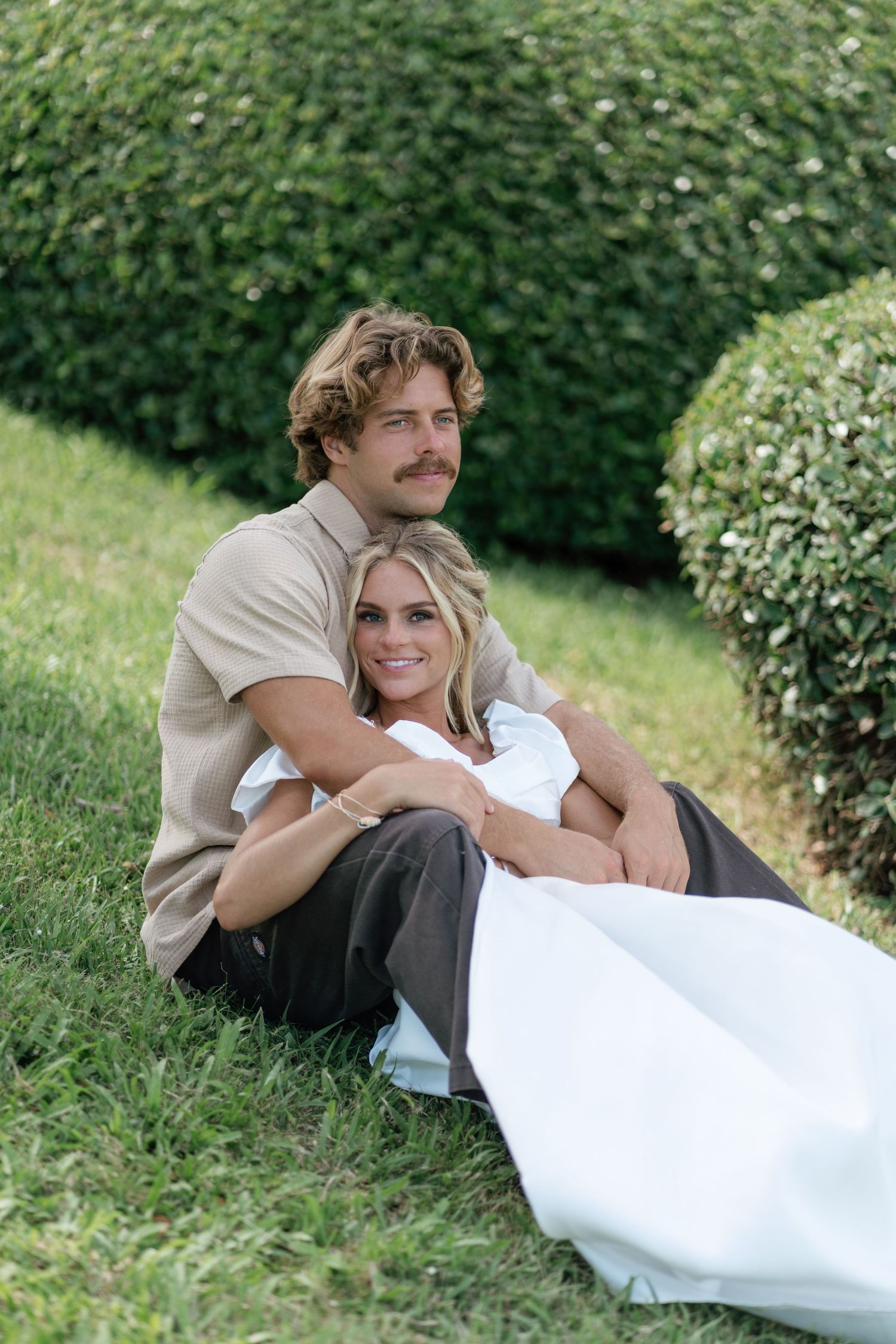 A couple sits together on green grass surrounded by manicured hedges, the woman leaning back against the man.