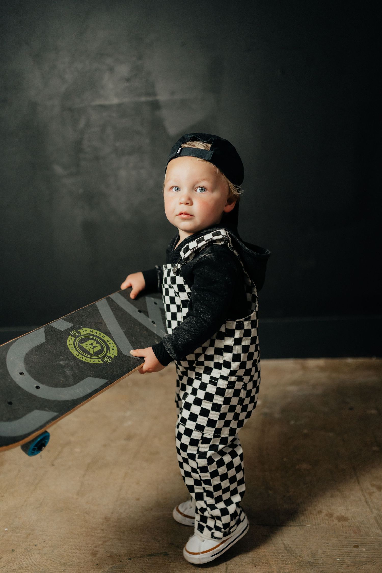 Toddler in checkered overalls and cap holding a skateboard, posing against a dark gray wall.